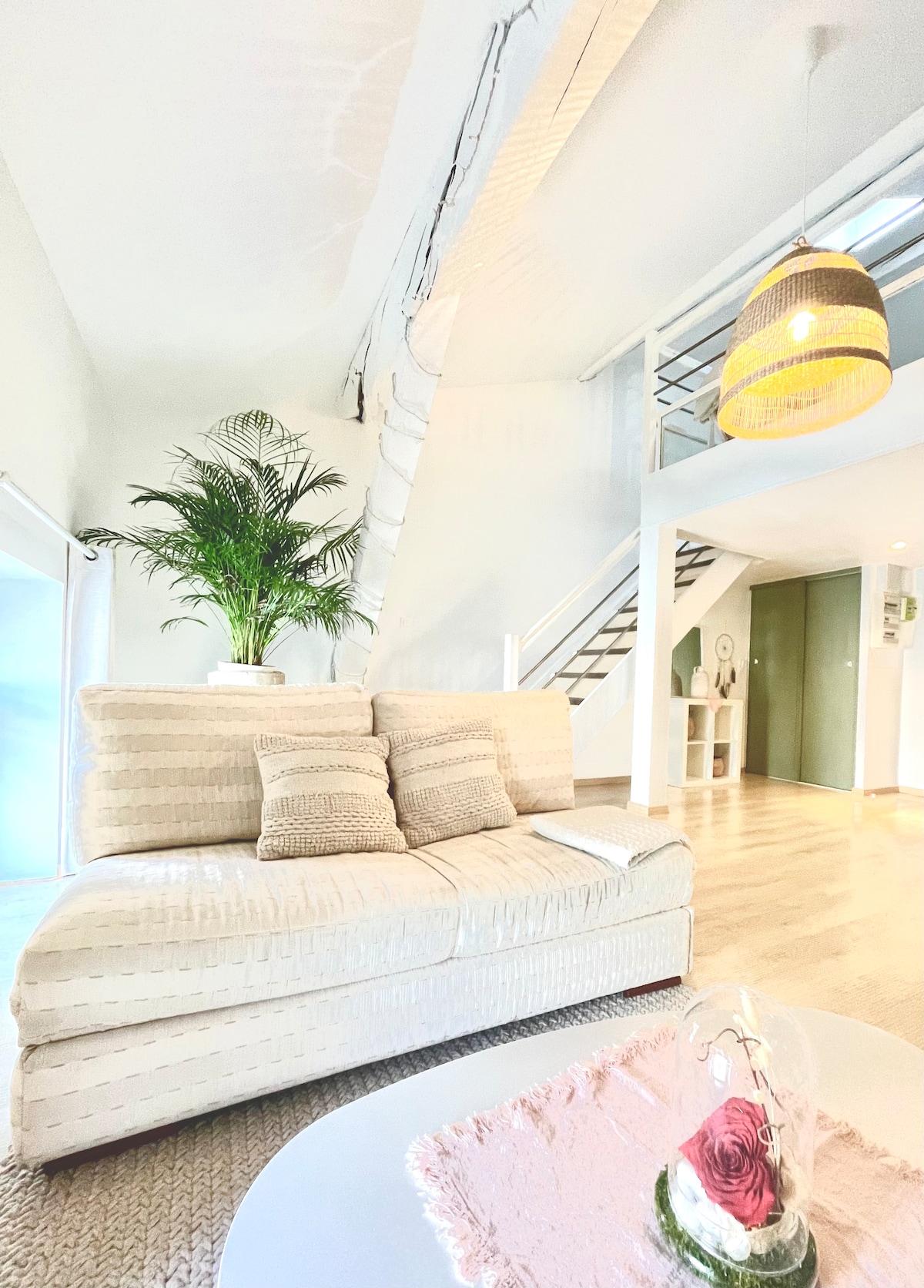 A stylish living area features a light-colored sofa with textured pillows, complemented by a modern pendant light overhead. A vibrant green potted plant adds a touch of nature. A staircase in the background leads to the mezzanine level, enhancing the open and airy feeling.
