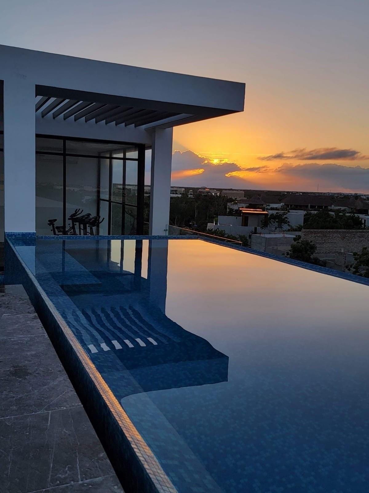 An infinity pool reflects the vibrant colors of the sunset, creating a serene atmosphere. The rooftop setting provides a panoramic view of the surrounding area, with fitness equipment visible in the background, offering a blend of leisure and activity.