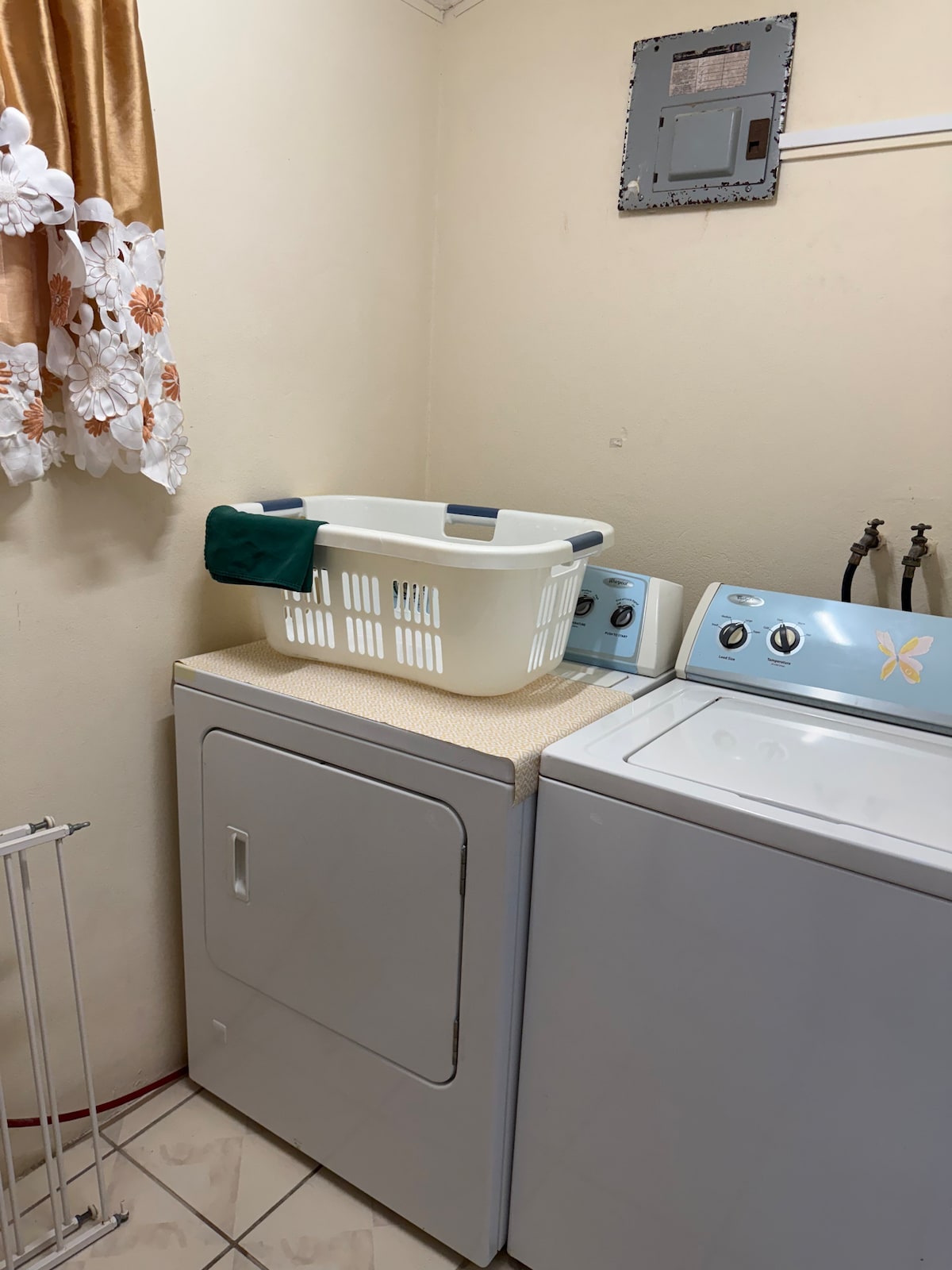 A laundry area is presented, featuring a stacked washing machine and dryer with a light blue exterior. A white laundry basket is placed on top of the dryer, accompanied by a green cloth. The space is well-lit, with beige walls and floral curtains.