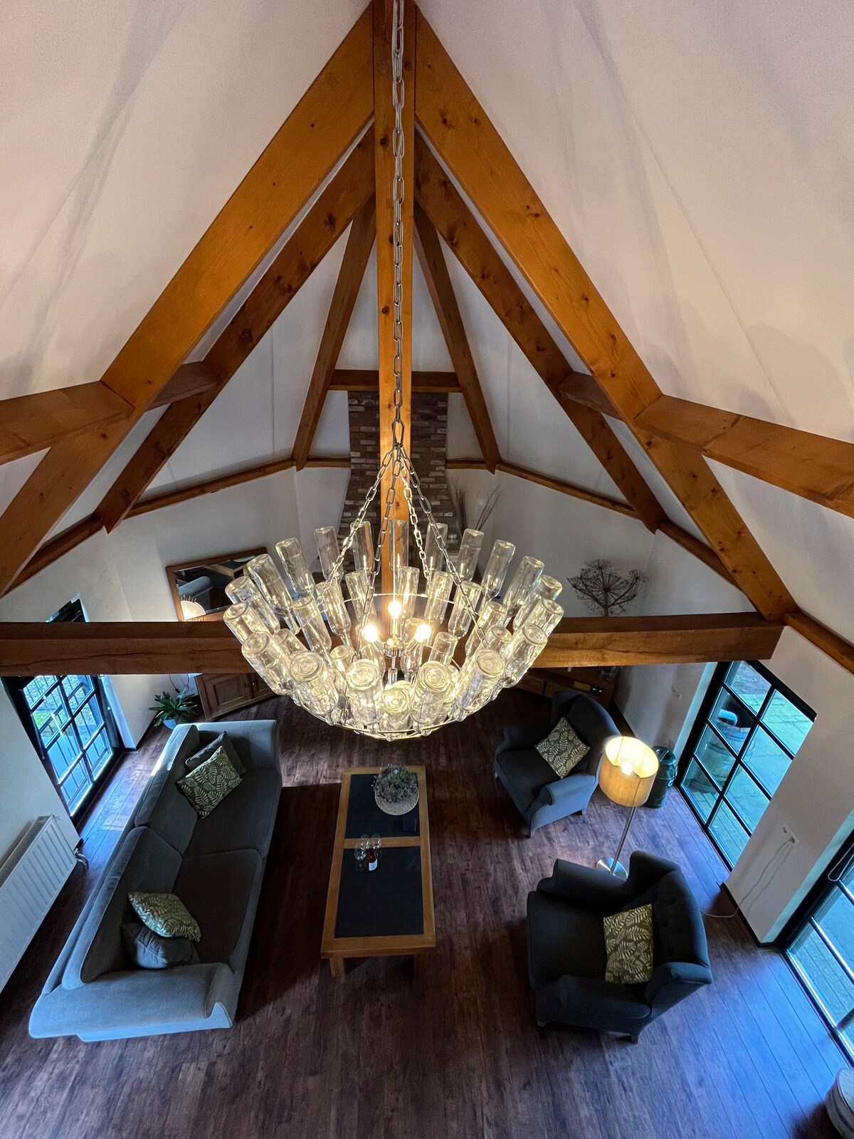 A spacious living area is highlighted by a striking chandelier hanging from a high, wooden-beamed ceiling. Two comfortable sofas with decorative cushions face a central table, while large glass doors lead to a patio, allowing natural light to fill the room.