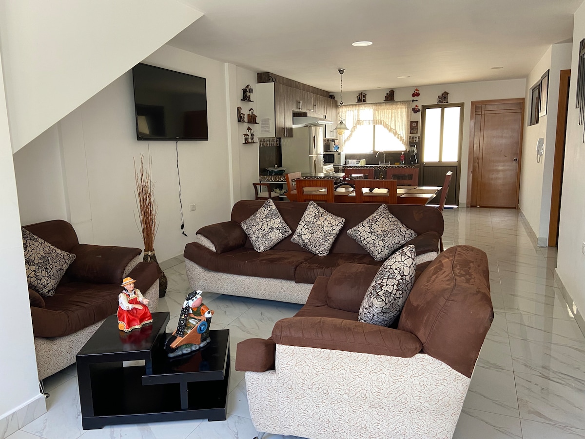 A living area is presented with two comfortable brown sofas adorned with decorative cushions and a coffee table in the center. A television is mounted on the wall, while a dining area with wooden chairs and a table is visible in the background, with natural light filtering through a window.