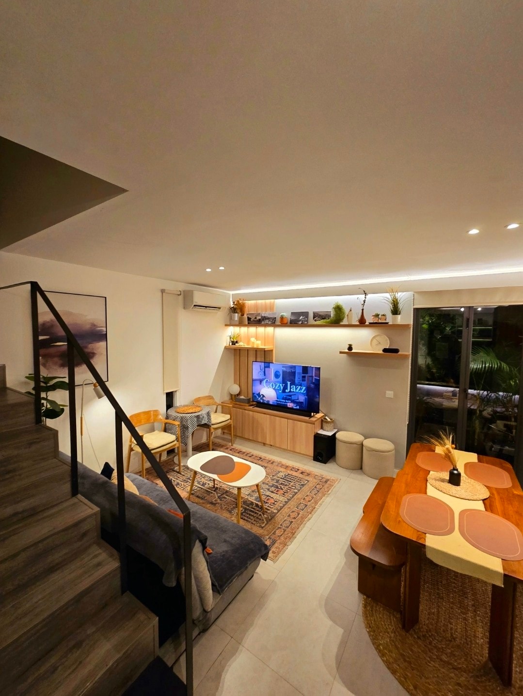 A spacious living area is shown with wooden accents and modern decor. A Sony Bravia TV is prominently displayed, surrounded by a cozy seating arrangement that includes a plush sofa and two chairs. A dining table for four, complemented by decorative items, is visible near the window.