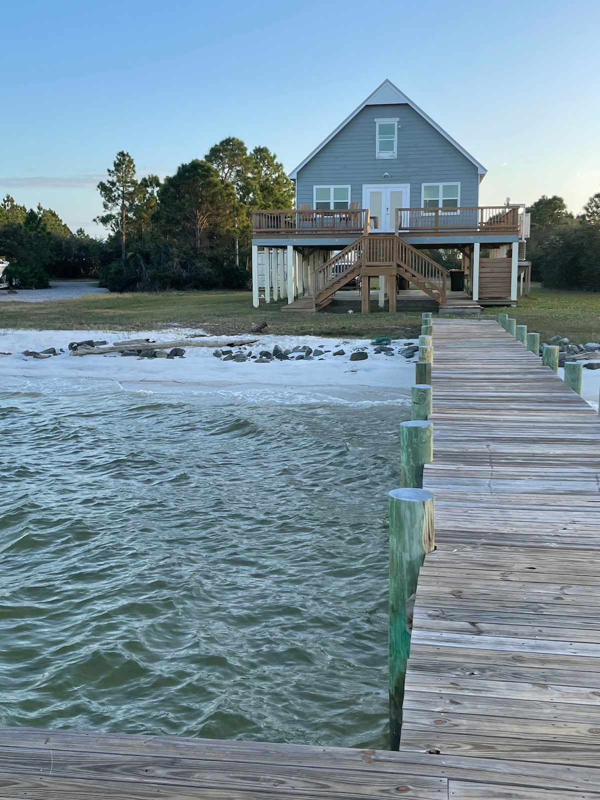 A wooden dock extends over the calm water, leading to a two-story beach house. The house is painted in soft grey with white trim, situated amidst trees. The sandy beach is visible, with gentle waves lapping at the shore.