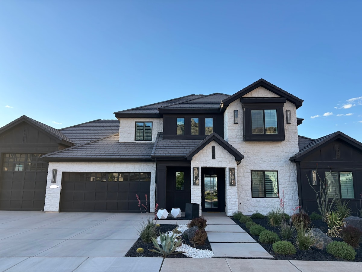 The exterior of the modern home displays a combination of white stone and dark accents, featuring multiple large windows. A clean, paved walkway leads to the entrance, surrounded by well-maintained landscaping with various shrubs and decorative elements.