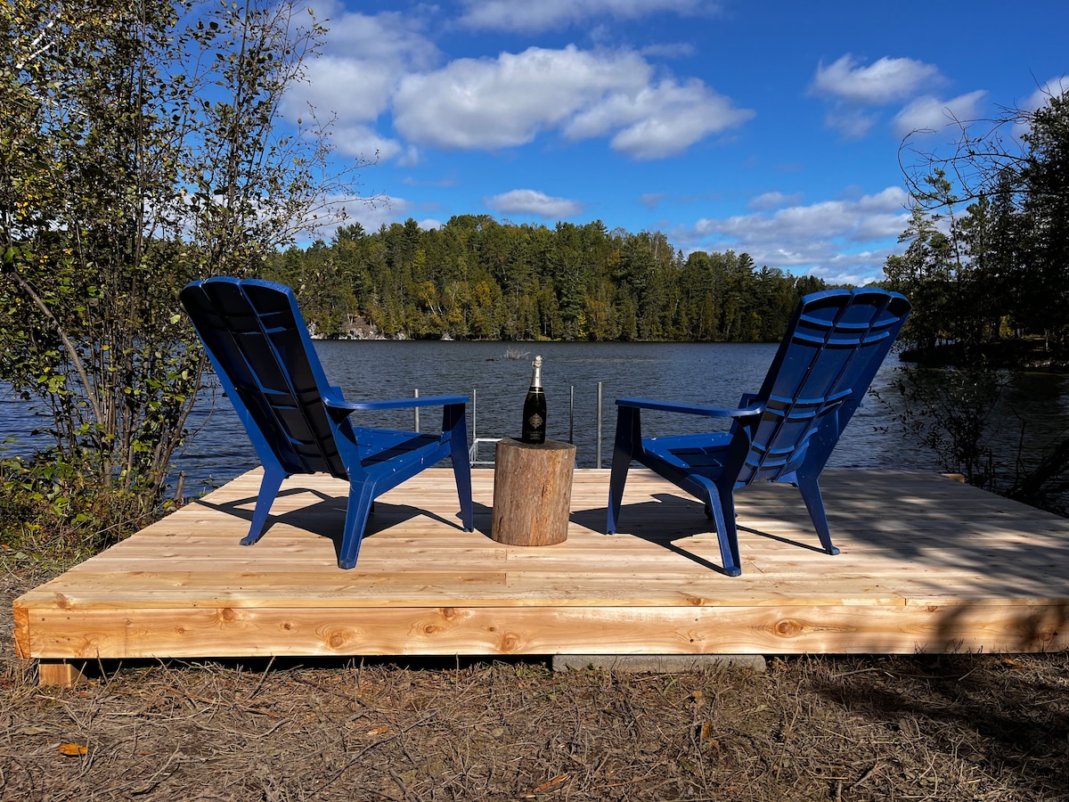 A wooden dock extends over the calm lake, featuring two blue Adirondack chairs positioned opposite each other. A small log serves as a table between them, and a bottle stands upright, creating an inviting space for relaxation amidst a natural setting.