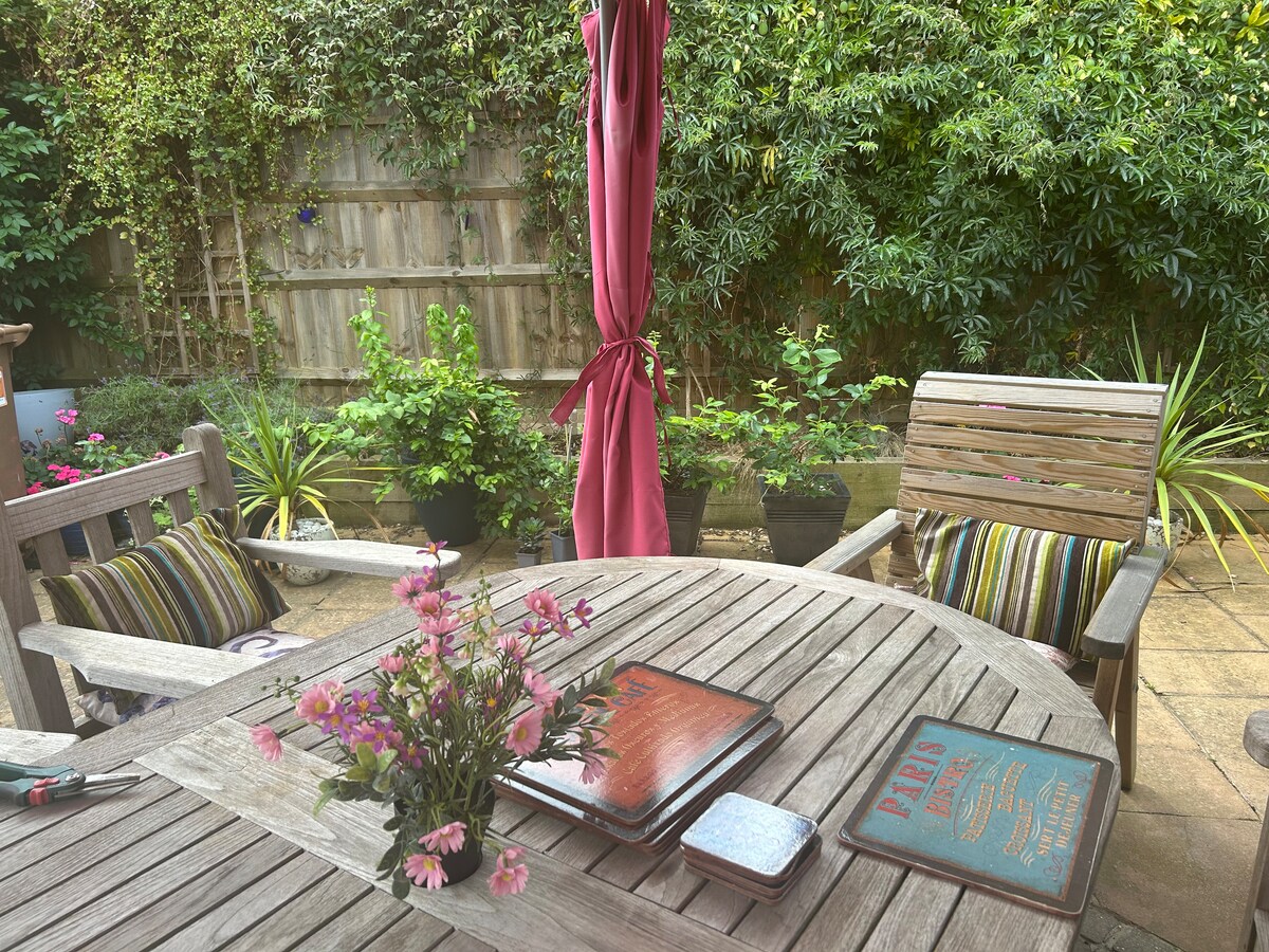 An outdoor patio area is showcased, featuring a wooden table surrounded by wooden chairs with striped cushions. A small vase of flowers is positioned on the table. Lush greenery including potted plants creates a serene backdrop, enhancing the environment's peacefulness.