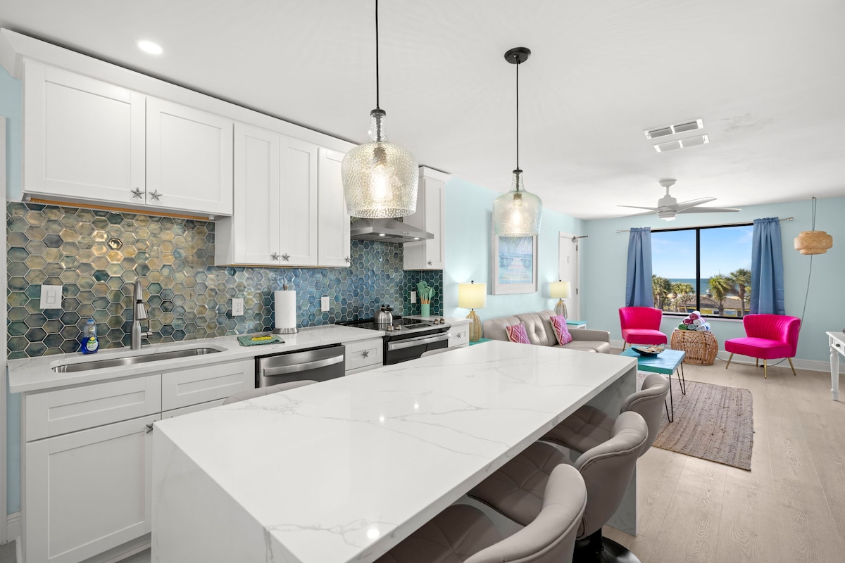 An open kitchen area displays modern white cabinetry and a marble countertop with seating. The backsplash features textured blue tiles, and pendant lights hang above. The adjoining living space is visible, featuring colorful chairs and large windows that allow natural light to brighten the room.