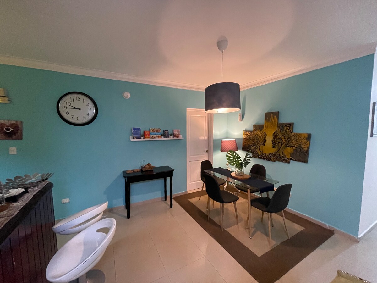 A dining area with a round table surrounded by four black chairs sits on a beige rug. A mural depicting yellow leaves is positioned on the wall, complemented by blue walls. A clock and decorative items are displayed on a shelf nearby, along with a small desk in the corner.