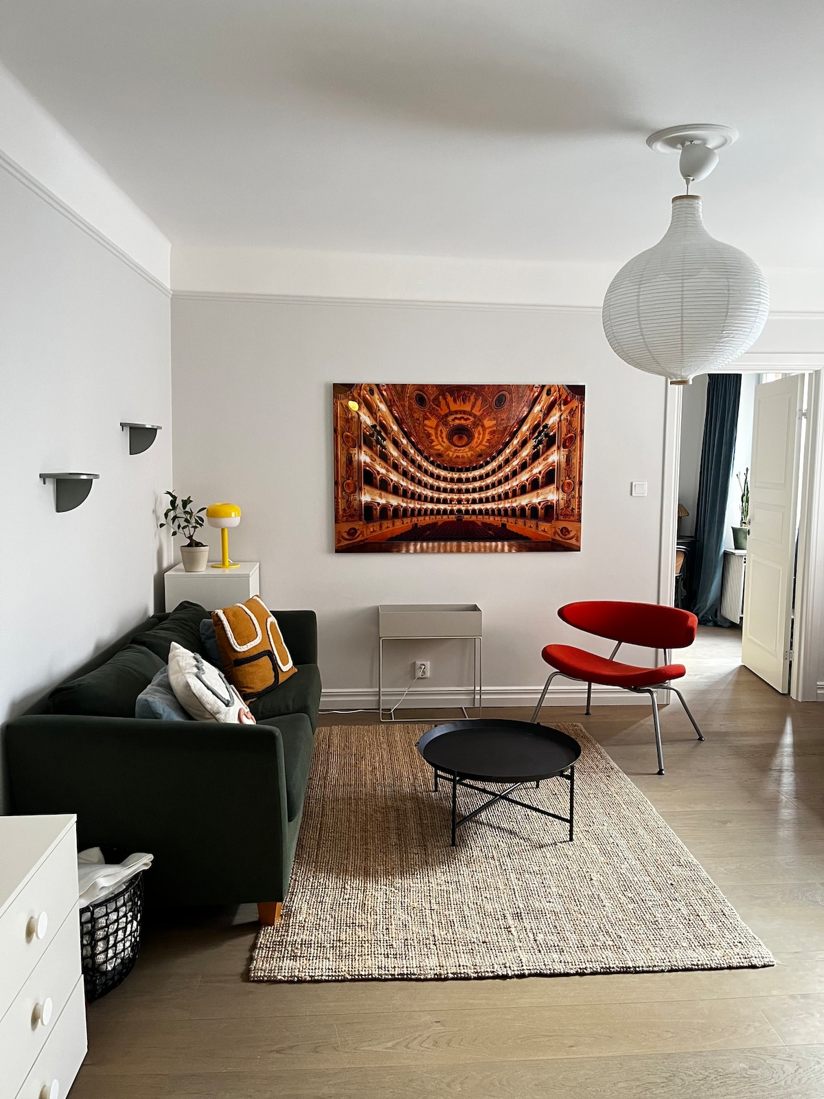 A modern living area features a green sofa paired with a bright red accent chair. A round coffee table sits atop a textured area rug, complemented by an artistic wall hanging. Natural light enters through an open doorway, illuminating the space.