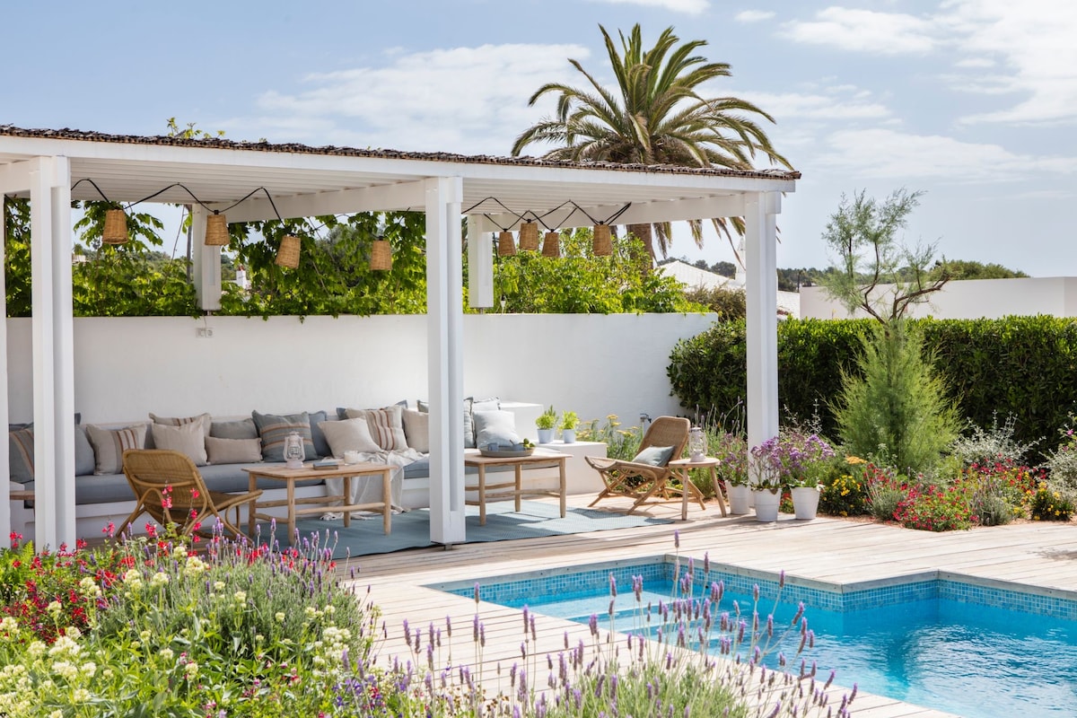 An outdoor patio area features a comfortable seating arrangement under a shaded roof, complemented by decorative hanging lights. Lush greenery and flowering plants border a tranquil swimming pool, providing a serene environment perfect for relaxation.