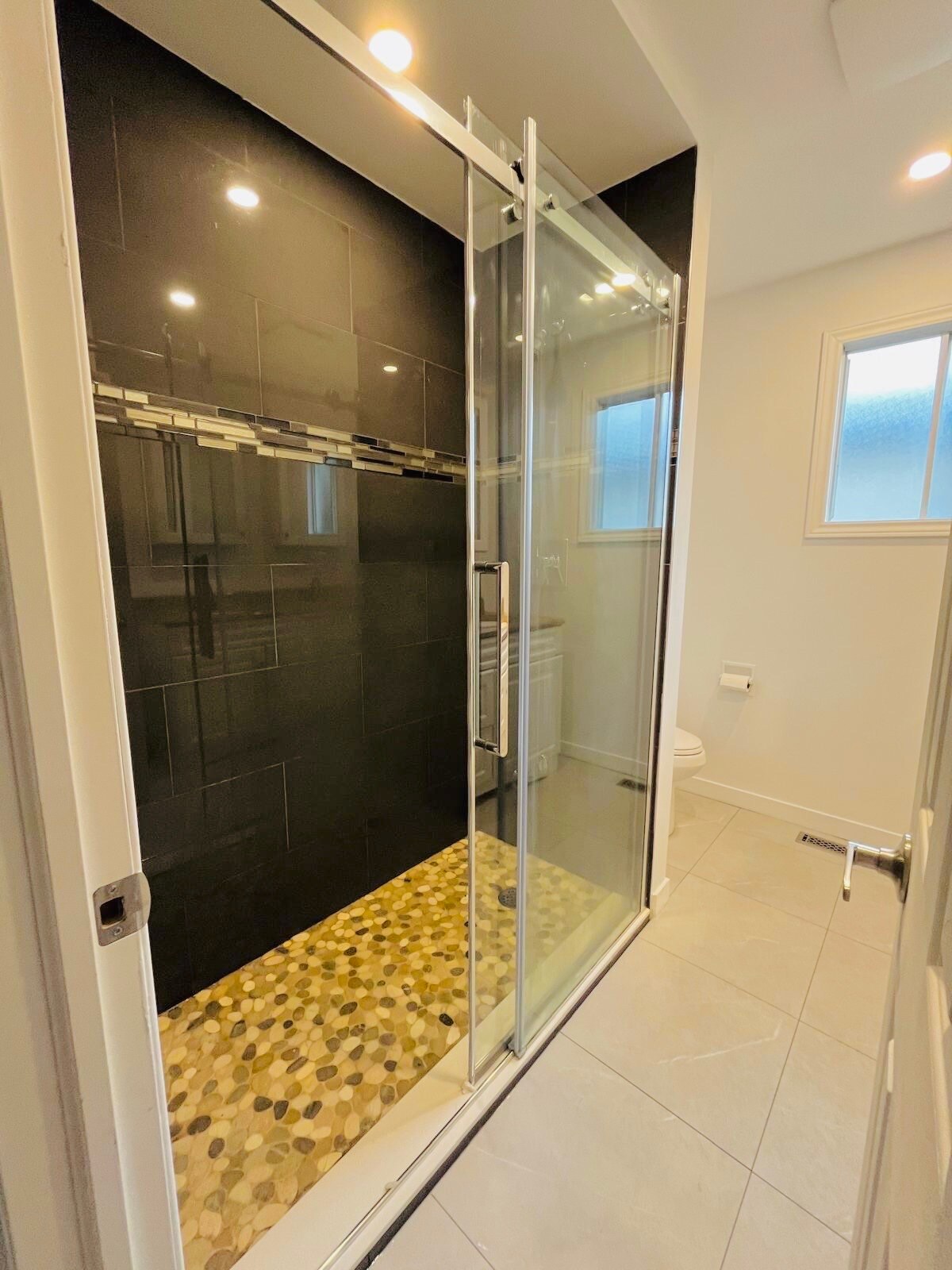 A modern bathroom is displayed, featuring a glass-enclosed shower with black tiles and a pebble stone floor. Natural light enters through a small window nearby, illuminating the space. A white toilet is located in the corner, complementing the light-colored tile flooring.