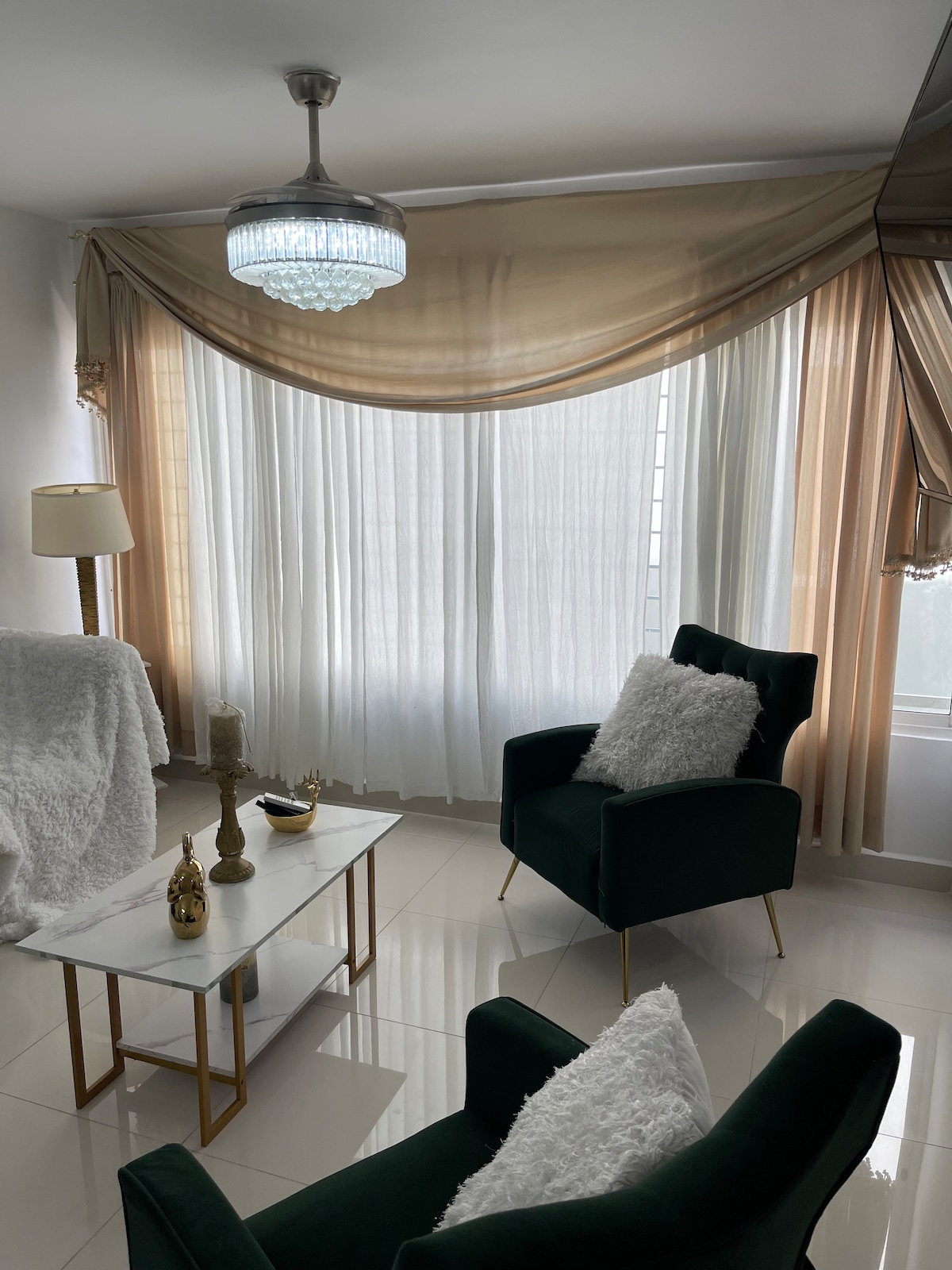 A well-lit corner of the living area is highlighted by soft natural light filtering through sheer curtains. Two plush green armchairs are positioned beside a marble-top coffee table adorned with decorative items. A stylish light fixture hangs from the ceiling, enhancing the welcoming ambiance.