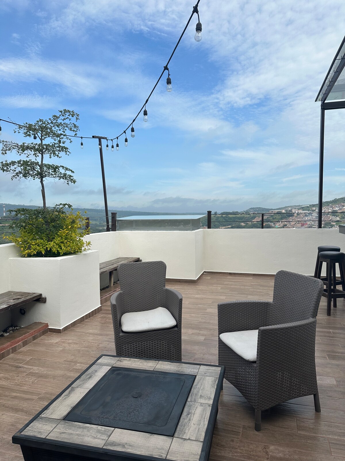 A spacious terrace features a modern seating area with two armchairs and a low table. Decorative plants and string lighting add a cozy touch, while panoramic city views are visible in the background under a clear blue sky.