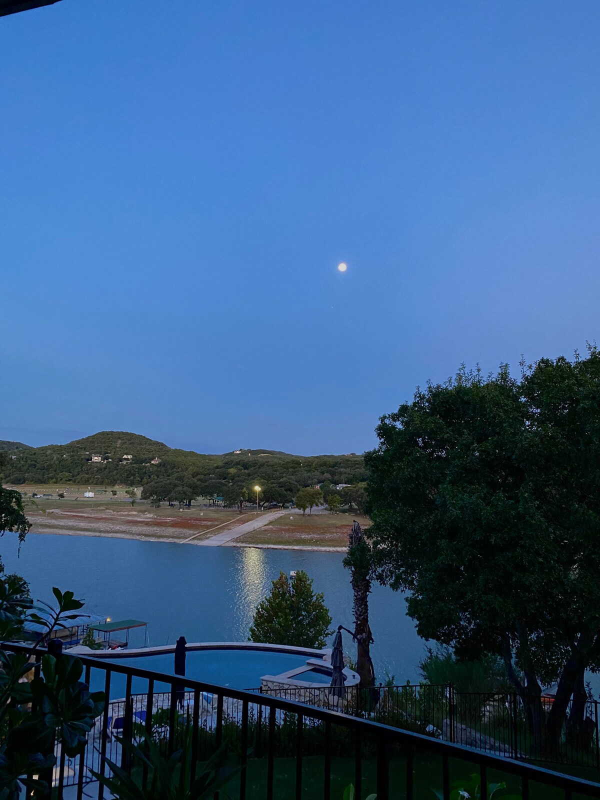 A serene view features a calm body of water beneath a fading twilight sky, with the moon visible. Gentle hills are seen in the background, accentuated by soft lighting from the shore. The image presents a tranquil atmosphere reflecting the natural surroundings.