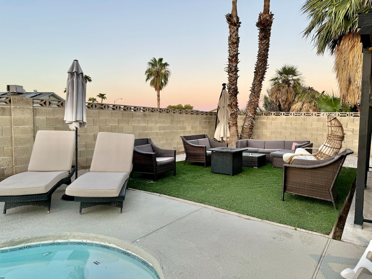 A well-appointed outdoor space features two lounge chairs and various seating options, including a shaded area with an umbrella. Palm trees provide a touch of greenery, while a hot tub is located nearby on the patio, creating a relaxing environment.