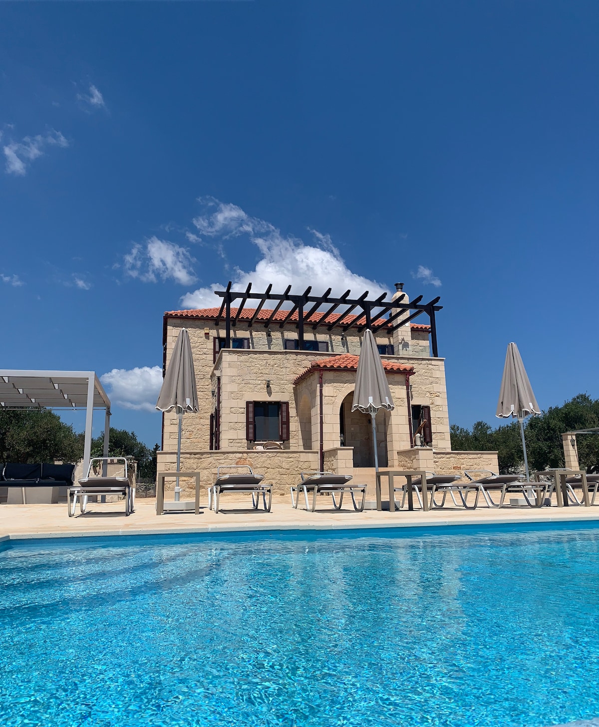 A stone villa with a classic design is showcased against a clear blue sky. The exterior features a shaded terrace with several sun loungers and umbrellas, while a pristine swimming pool reflects its charming façade.