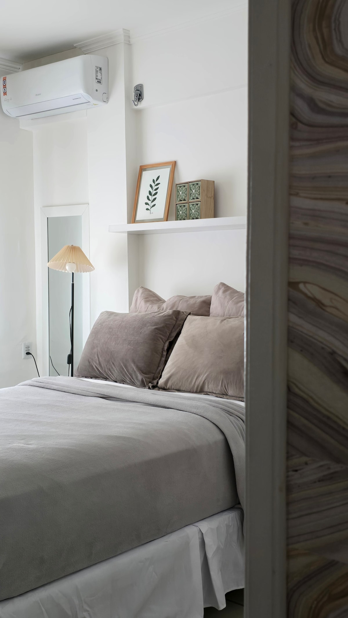 A serene bedroom setting is showcased with a soft-toned bed adorned with plush pillows. Natural light enters from an adjacent window, illuminating the space. A lamp and decorative wall art add subtle charm to the minimalist design. A full-length mirror is positioned nearby.