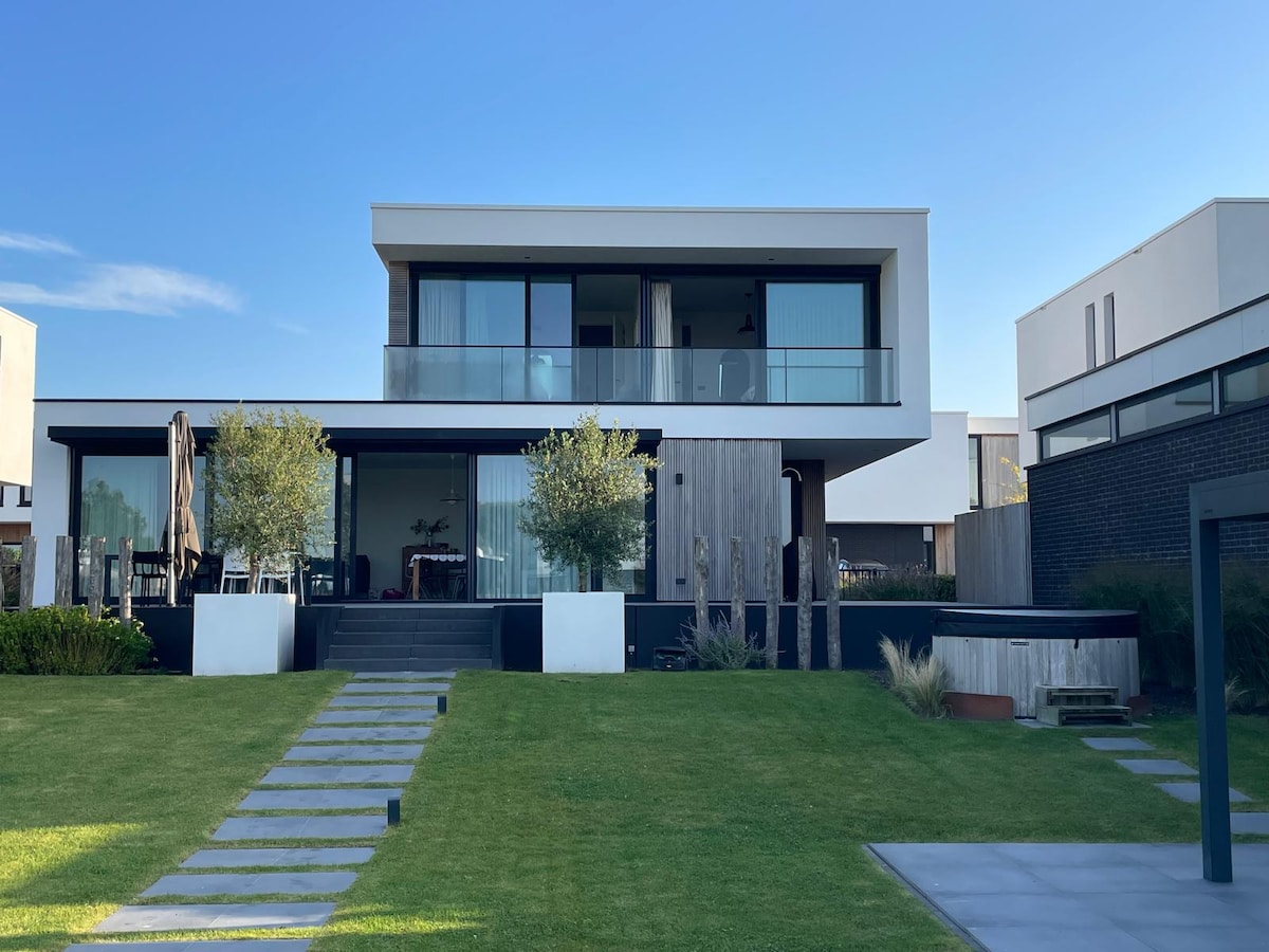 A modern two-story water villa is presented, featuring large glass windows and a spacious balcony. A well-manicured lawn leads to the entrance, lined with stone pathways. An electric hot tub is situated on the right, surrounded by greenery, contributing to the villa's outdoor appeal.