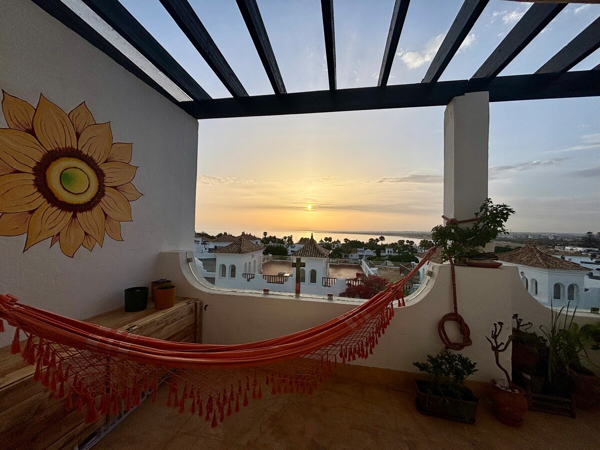 A spacious terrace features a vibrant mural of a sunflower, alongside a comfortable hammock. The sunset casts warm hues over the horizon, creating a serene view of the sea and surrounding landscape. Potted plants are arranged around the area, contributing to the inviting setting.