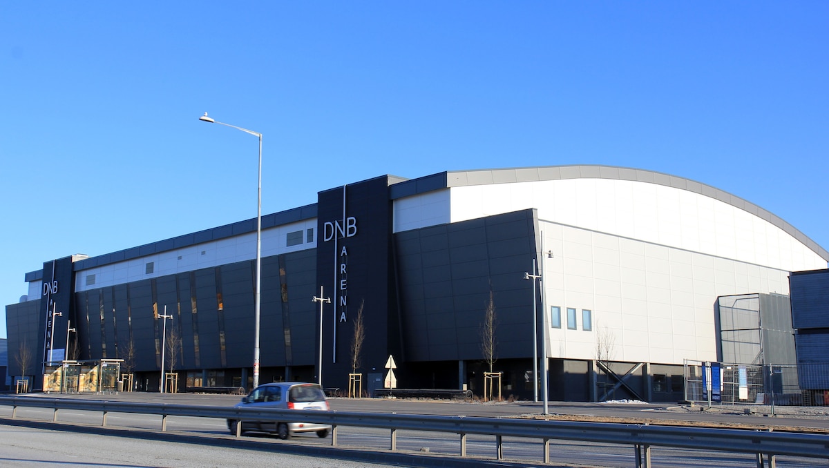 The DNB Arena is a large, modern building with a distinctive curved roof. The exterior features a combination of black and white panels, prominently displaying the arena's name. Nearby lighting poles and an open area are visible, with a road and parked cars in the foreground.