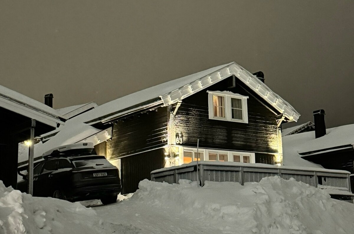 A charming black wooden cabin is illuminated by warm lights, nestled amidst snow-covered terrain. A sloped roof is blanketed in snow, and a car is parked nearby. Large windows provide a glimpse of the inviting interior, framed by a welcoming porch.