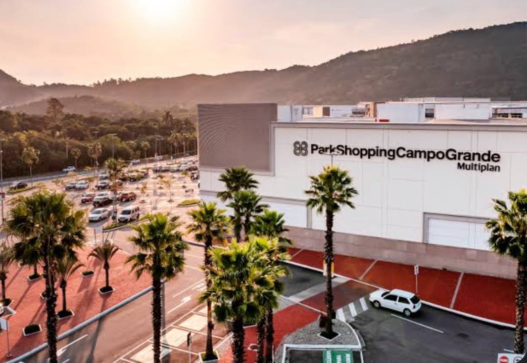 An aerial view captures the exterior of Park Shopping Campo Grande, showcasing the building's modern architecture surrounded by palm trees. The parking area is visible with vehicles and the surrounding landscape of mountains in the background, illuminated by the warm hues of sunset.