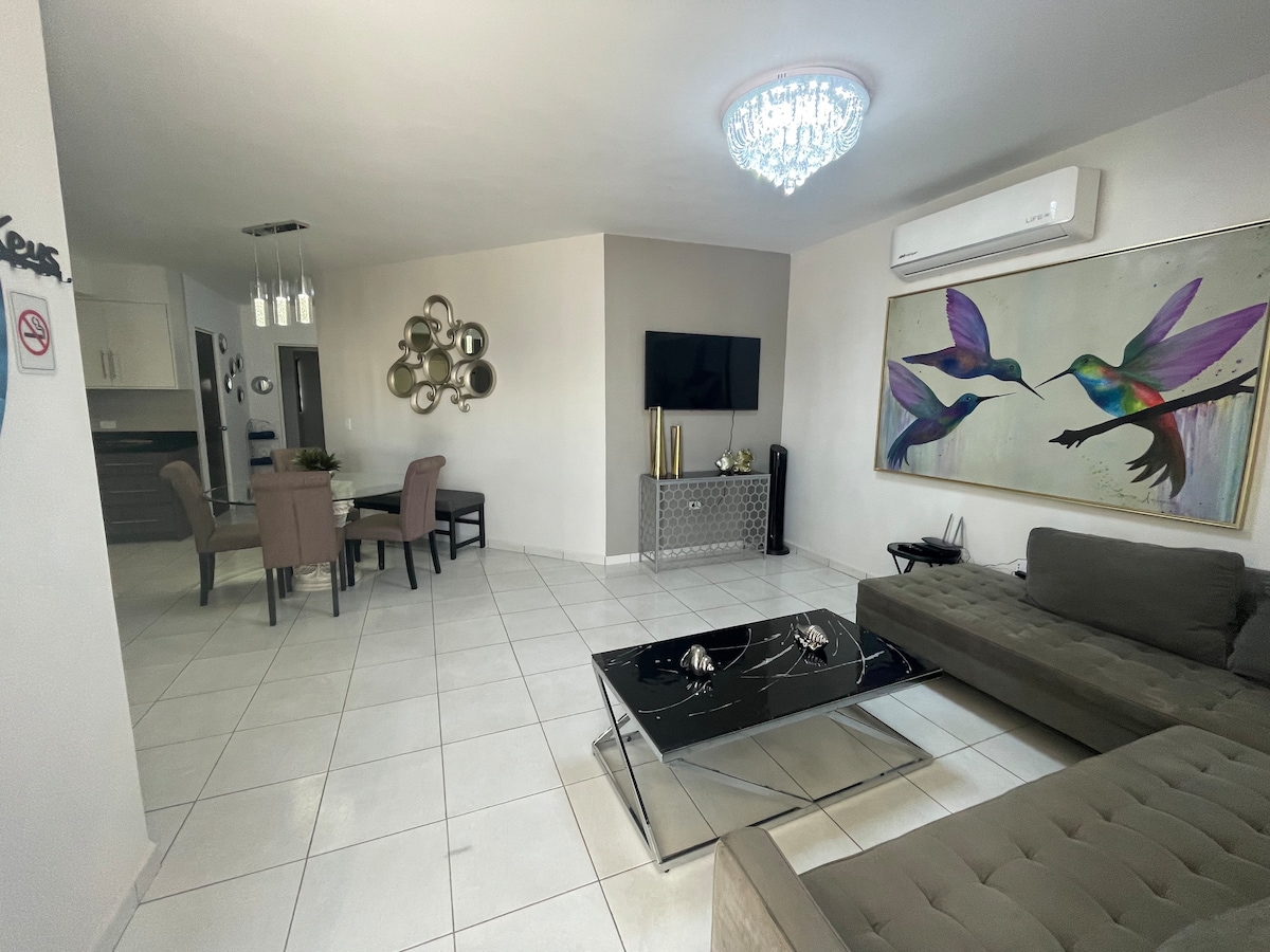 A modern living area features a stylish sectional sofa in a muted tone, complemented by a glass coffee table. A large artwork depicting colorful birds adorns the wall. An elegant chandelier provides illumination, while a TV is mounted against a softly colored wall.