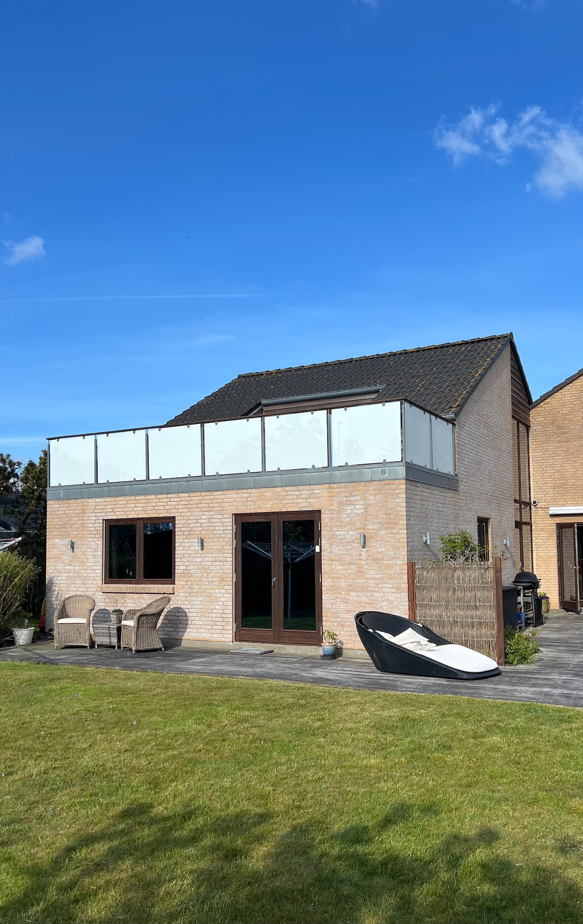 A well-designed exterior view of a light-colored villa is presented, featuring large glass-panel railings above the entrance. Comfortable outdoor seating is arranged on the patio, with a contemporary lounge chair positioned nearby. The green lawn adds a natural touch to the overall setting.