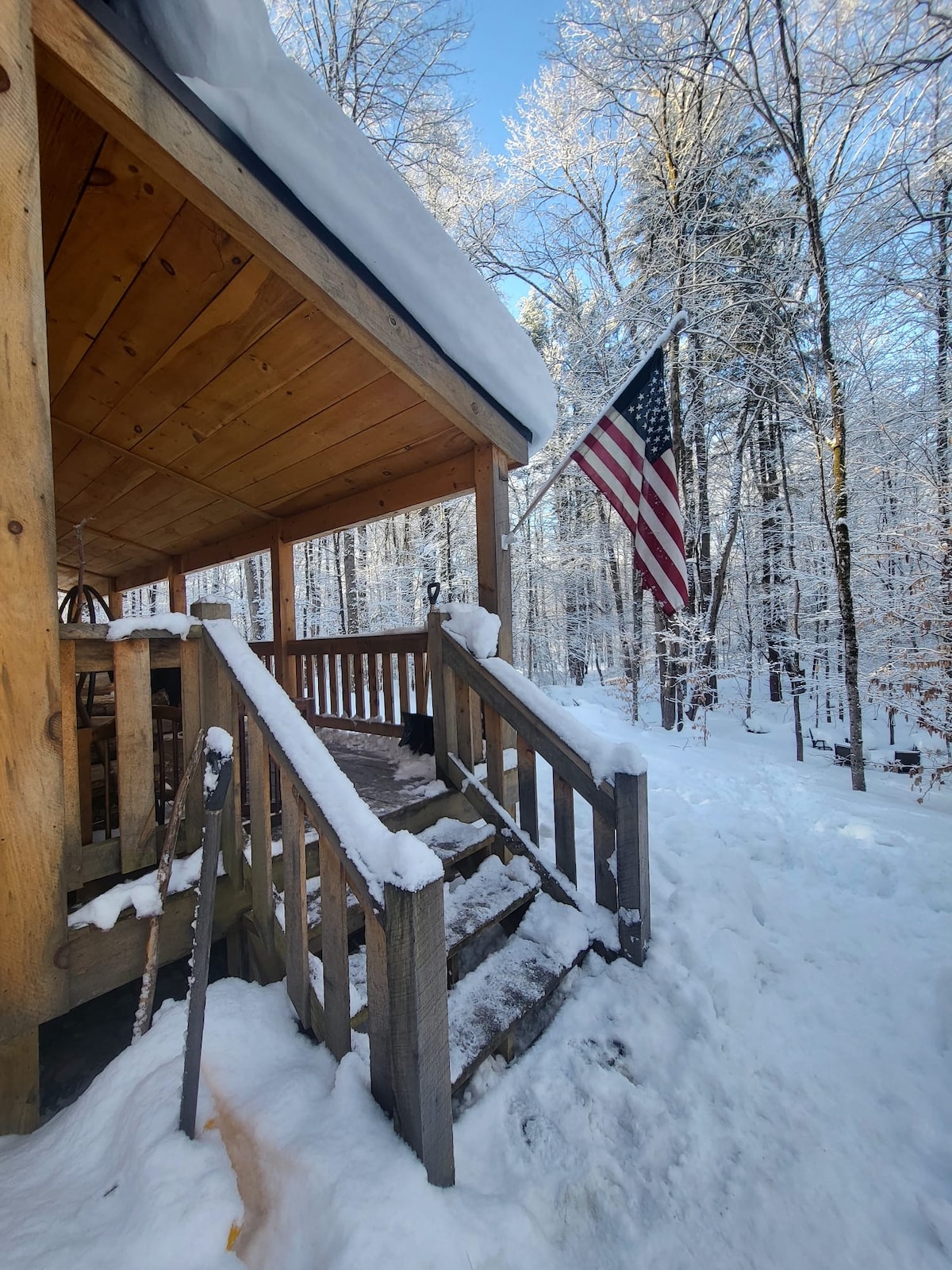 The entrance to the cabin is framed by a wooden porch, flanked by snowy steps. An American flag flutters in the crisp winter air, with snow-laden trees providing a serene backdrop. The scene evokes a tranquil, wintry atmosphere.