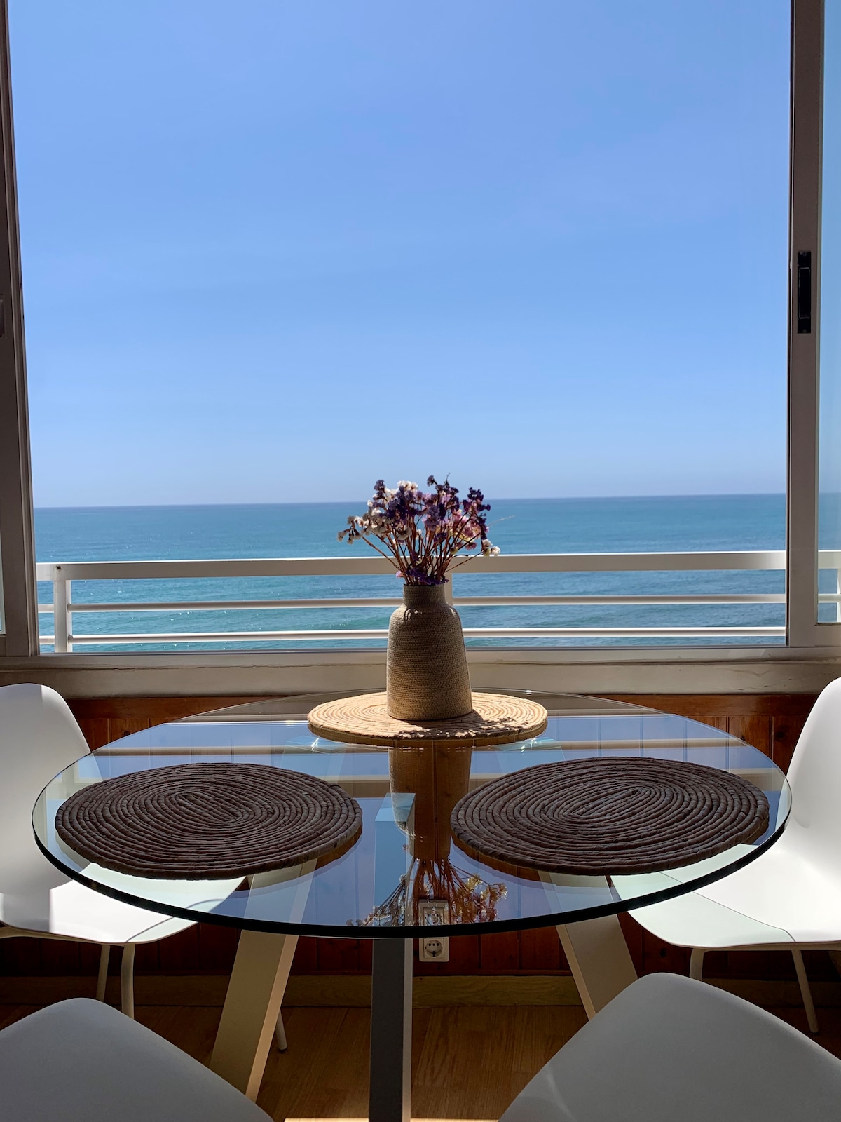 A glass dining table is set with natural woven placemats and a vase of dried flowers at its center. The area is framed by large windows offering a clear view of the sunny ocean and blue sky, creating a bright and airy atmosphere.