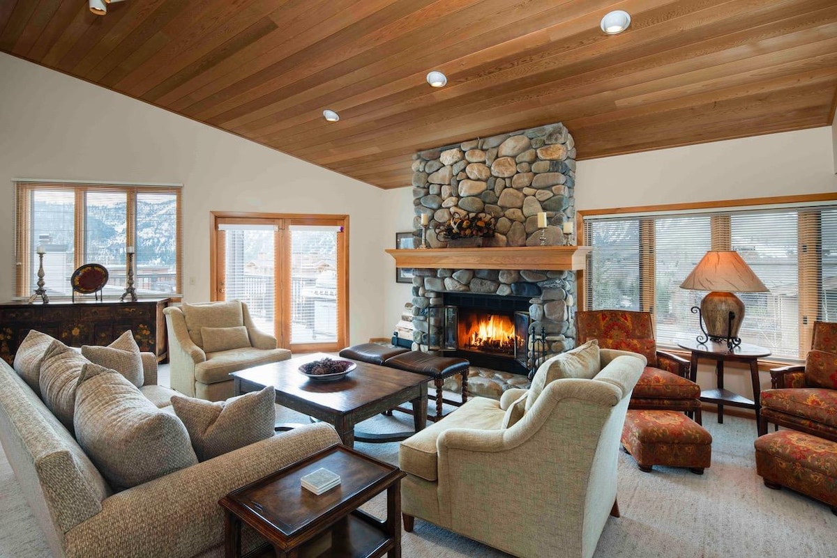 A spacious family room features a stone fireplace, creating a focal point within the warm wood-paneled space. Comfortable seating options are arranged around a central coffee table, complemented by large windows that allow natural light to fill the room.