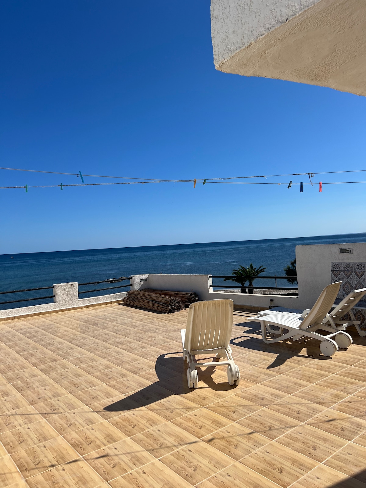 A spacious terrace extends towards the sea, featuring sun loungers positioned for relaxation. The wooden floor adds warmth to the area. A clear blue sky and calm waters create a serene backdrop, while colorful laundry lines stretch across, adding a touch of vibrancy to the scene.
