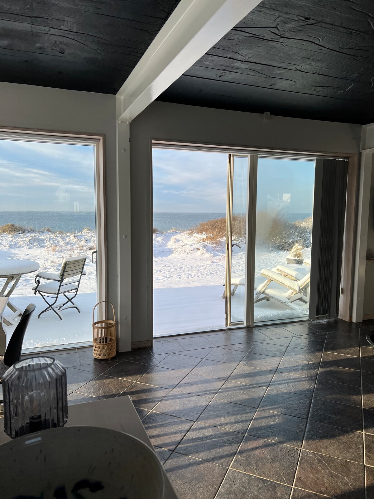 A bright open area showcases large glass doors that lead to a snowy outdoor deck with views of the sea. A dining table and chairs are positioned within the space, contrasting the sleek tile flooring and the dark ceiling above.