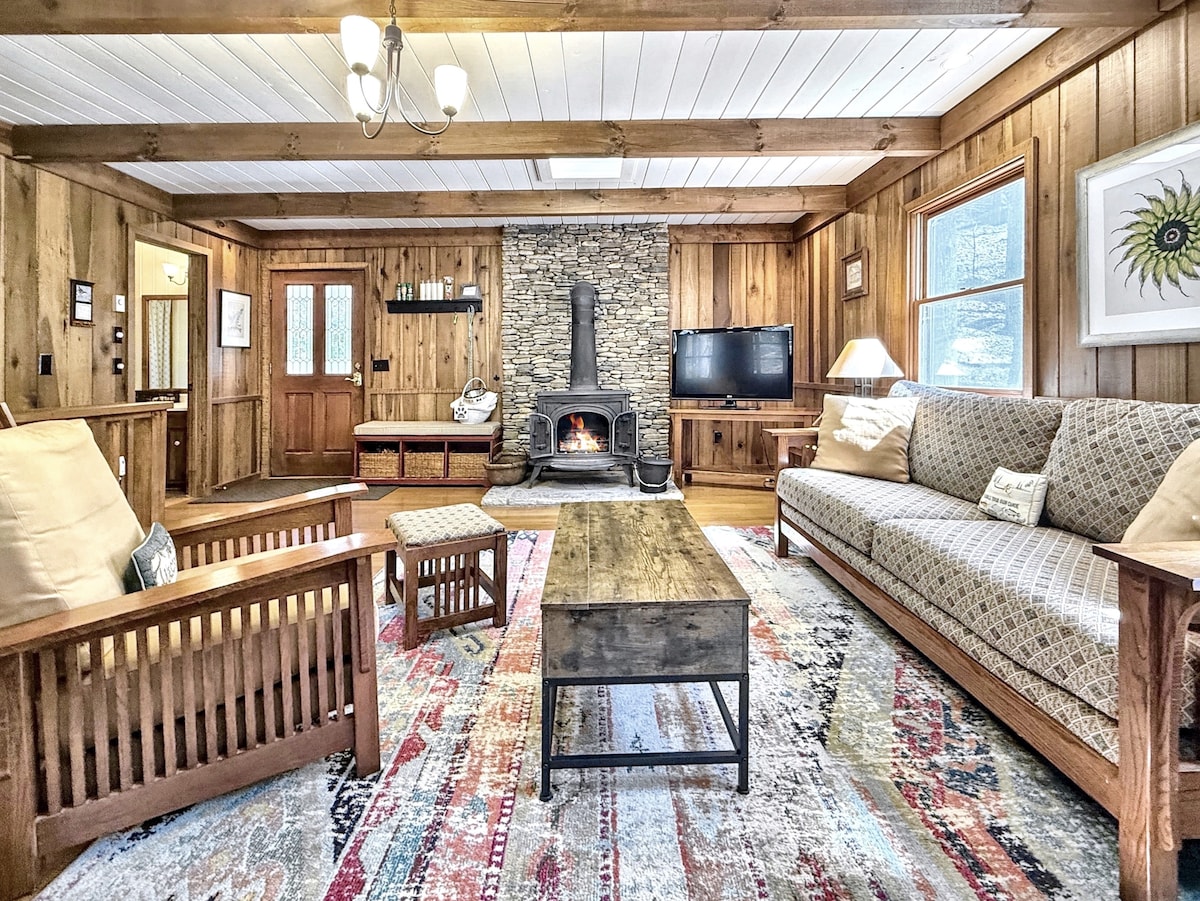 A cozy living area features natural wood paneling, a stone fireplace, and comfortable seating arrangements. A plush sofa and wooden chairs surround a rustic coffee table on a colorful area rug. Large windows invite natural light, enhancing the inviting ambiance.
