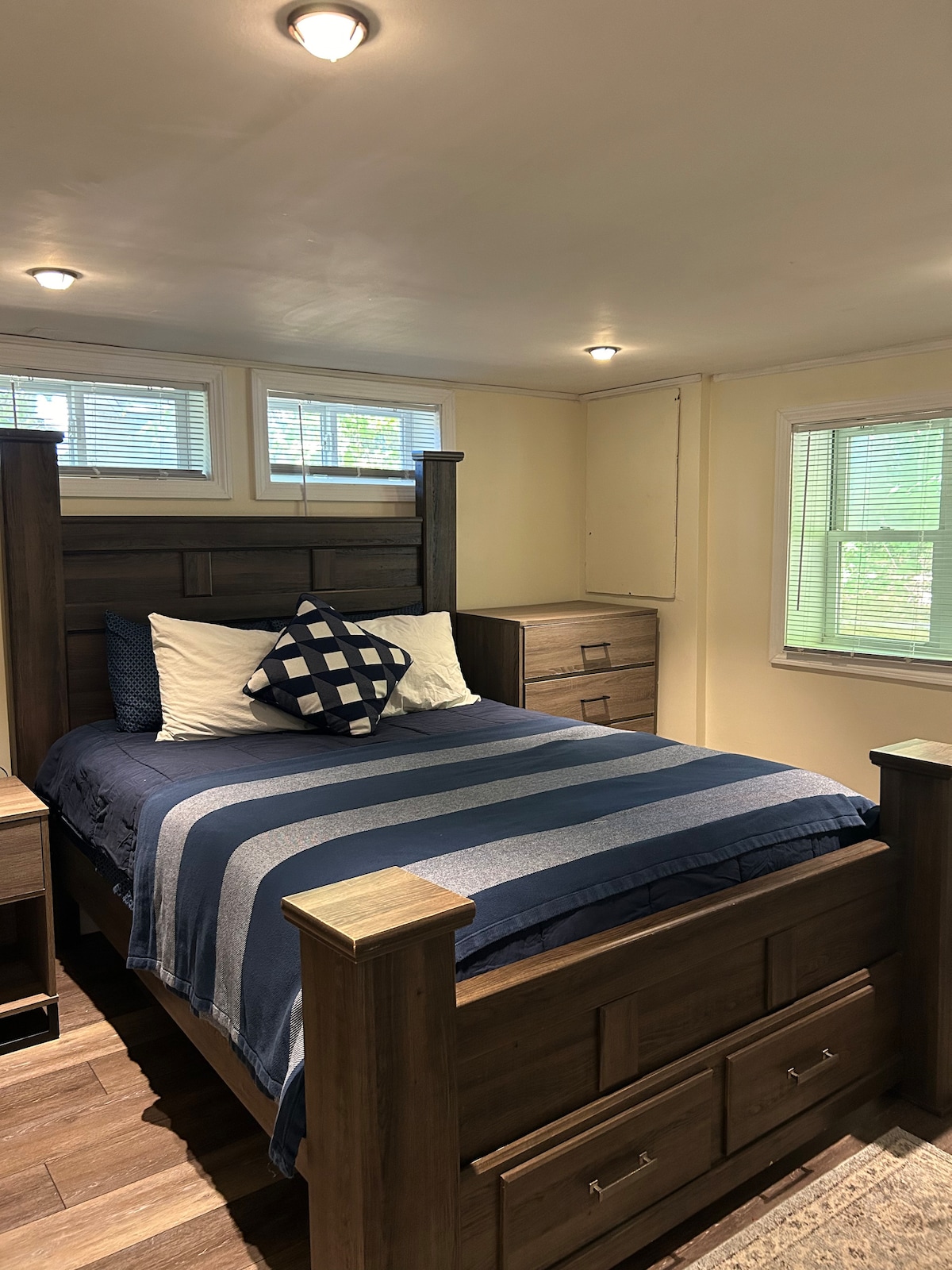A well-appointed bedroom is equipped with a spacious wooden bed featuring a dark blue and white striped blanket. Pillows with a geometric pattern are arranged at the head of the bed. Natural light enters through multiple small windows, illuminating the light-colored walls.