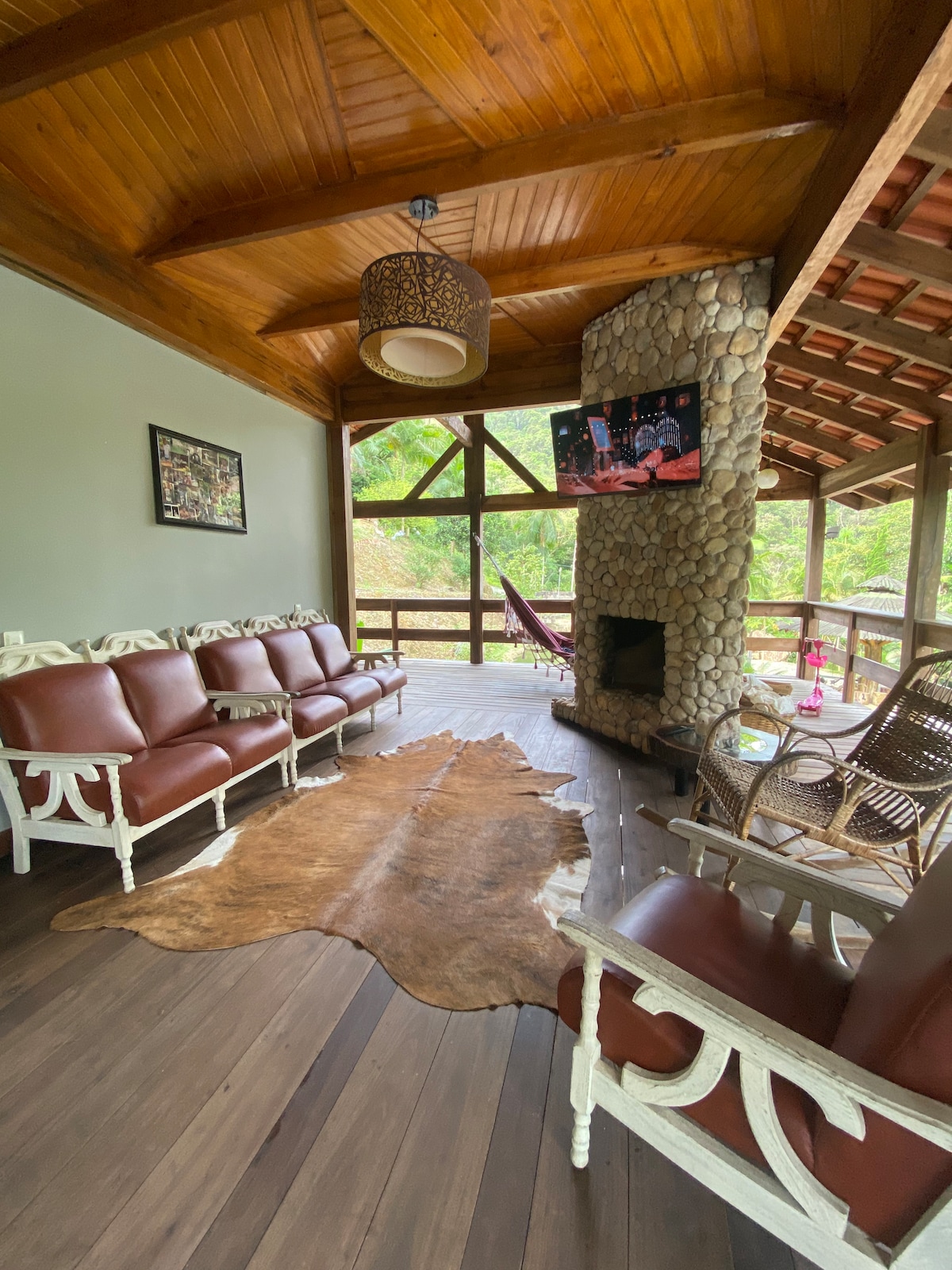 A spacious living area features comfortable brown seating arranged around a stone fireplace. A decorative light fixture hangs from a wooden ceiling. A flat-screen TV is mounted on the wall, with large windows offering views of the surrounding greenery.