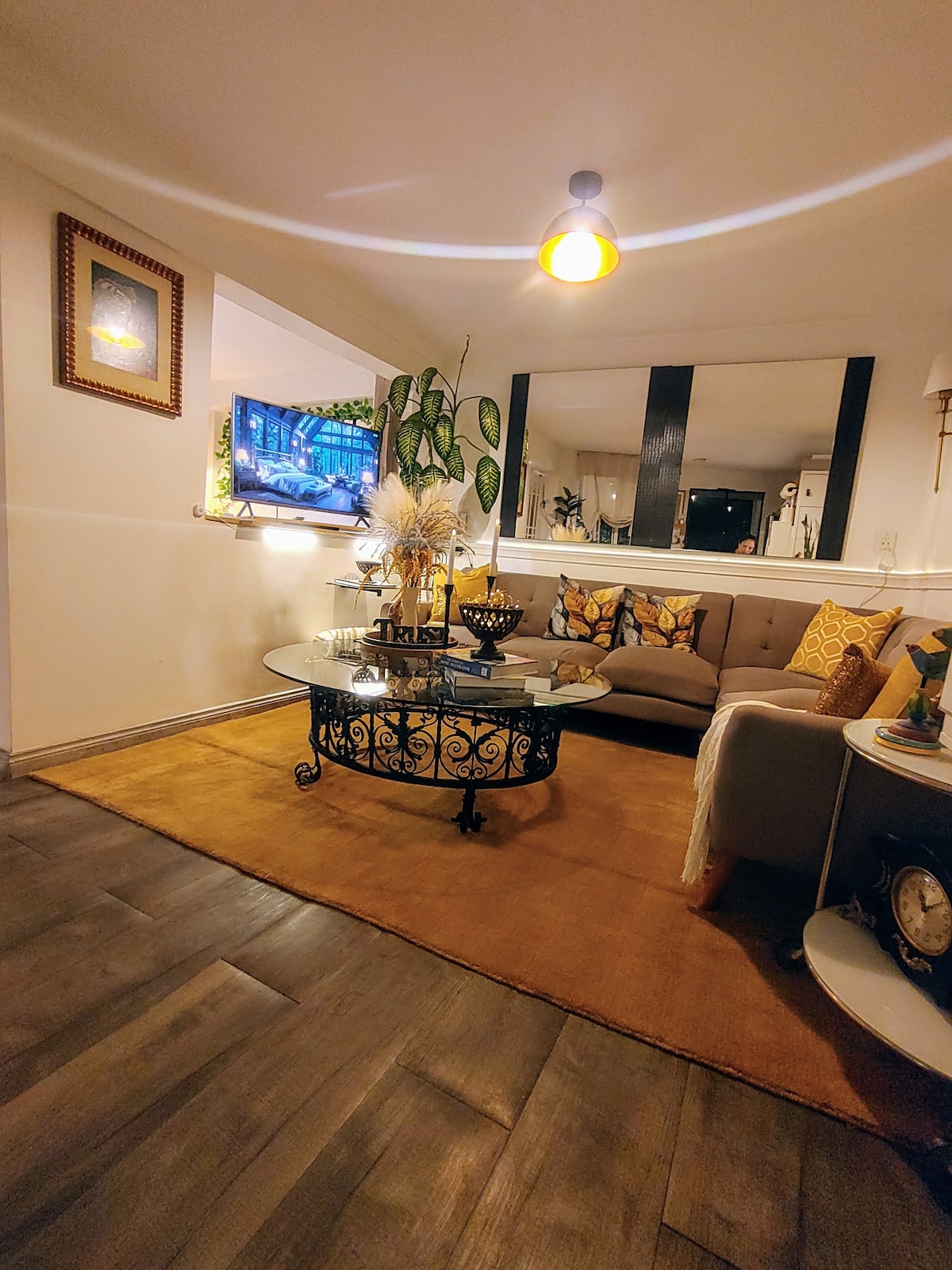A cozy living area features a comfortable sofa with decorative cushions, complemented by a round glass coffee table. A flat-screen television is mounted on the wall, surrounded by plants and framed artwork. Warm lighting is provided by a ceiling fixture, enhancing the space's inviting feel.