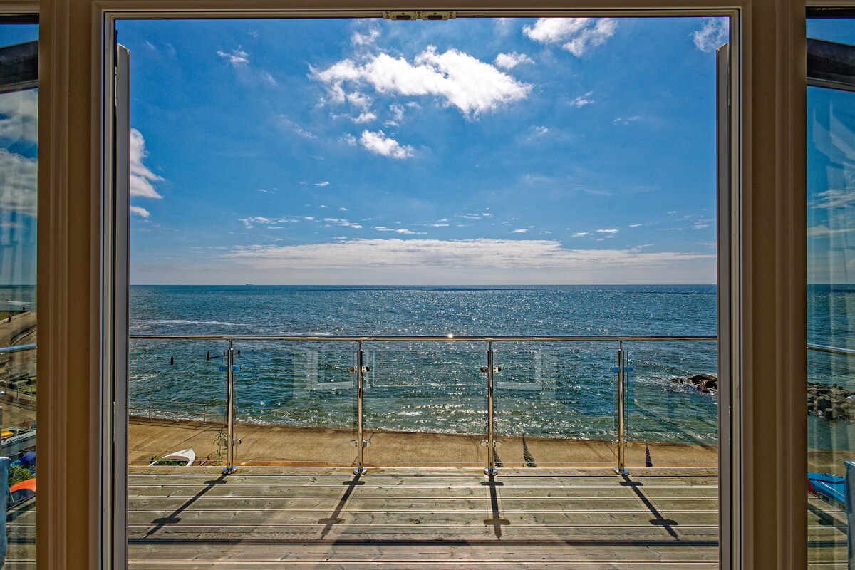 A large opening provides an unobstructed view of the sea, where gentle waves meet the sandy shore under a clear blue sky. A glass railing defines the outdoor space, enhancing the feeling of openness and connection to the coastal landscape.