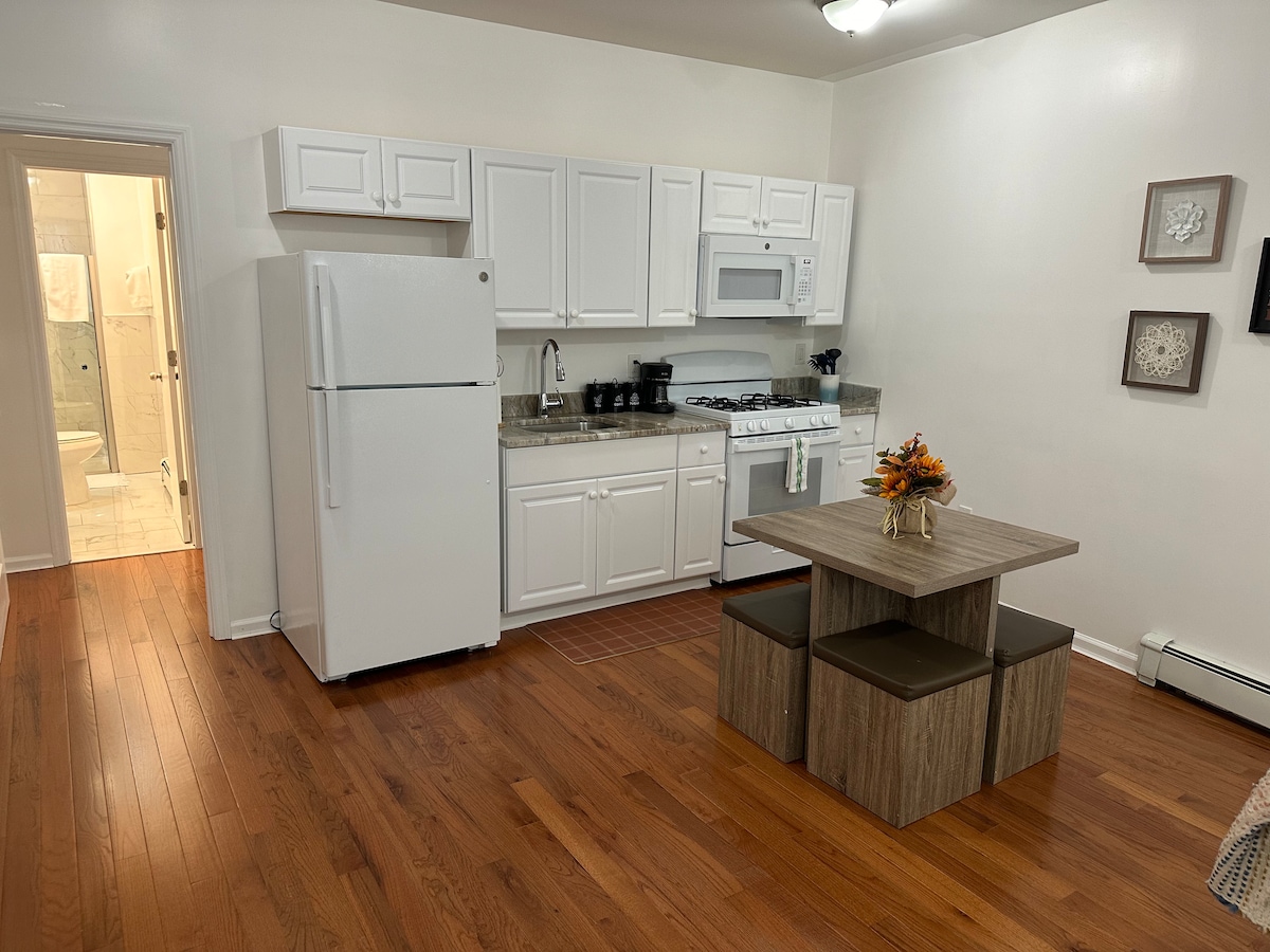 A compact kitchen area features white cabinetry and modern appliances, including a refrigerator and microwave. A dining table with two stools is positioned centrally, while the smooth wooden floor adds warmth. A glimpse of the bathroom is visible through a doorway at the back.