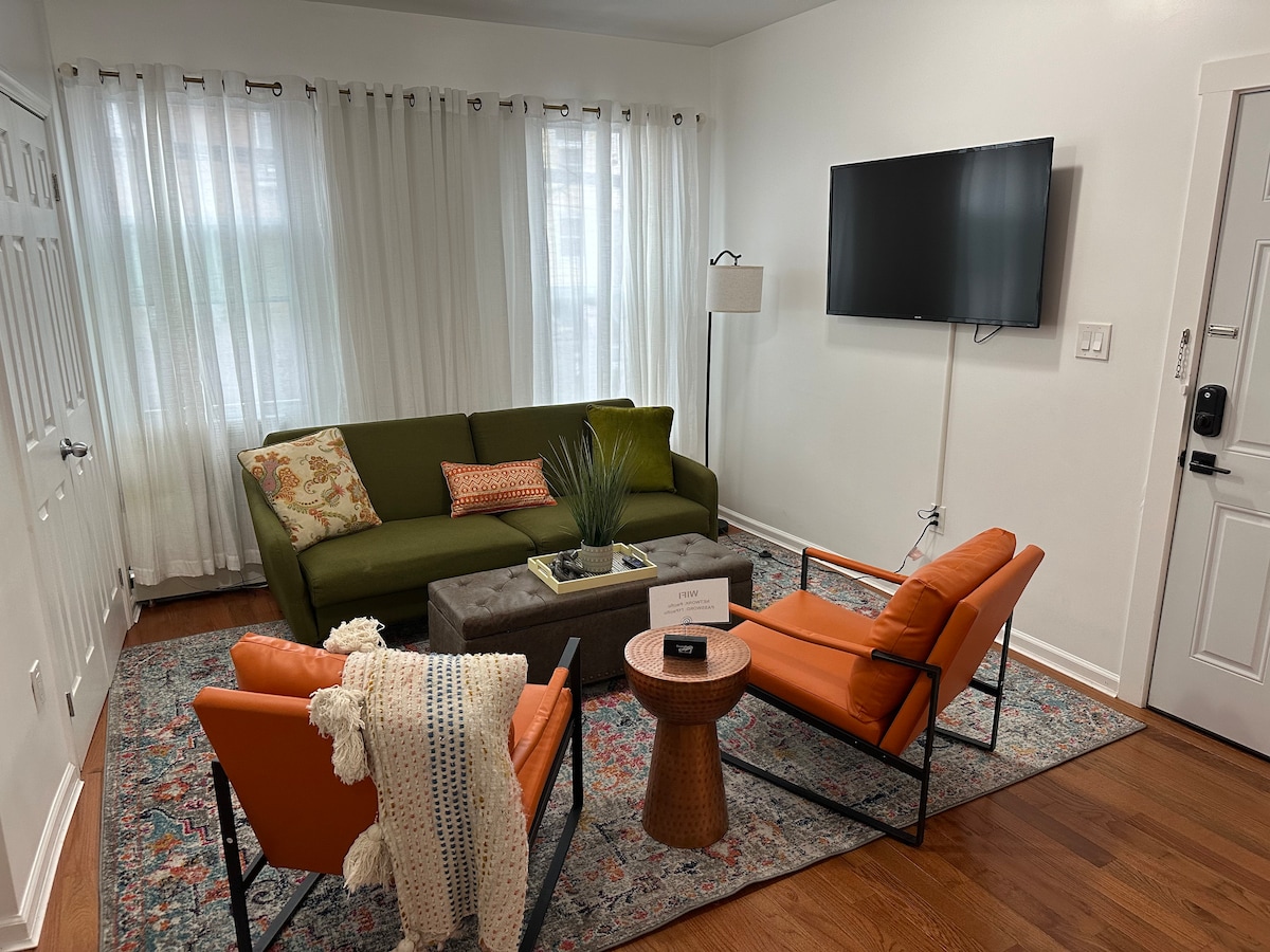 A comfortable living area features a green sofa adorned with decorative pillows. Two orange armchairs flank a wooden side table, which holds a small decor piece. Natural light filters through sheer white curtains, enhancing the warm ambiance. A wall-mounted television is visible.
