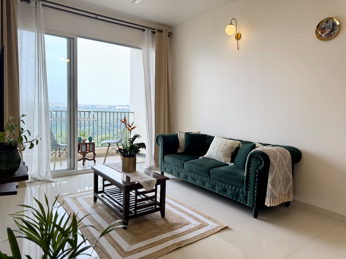 A cozy living area is highlighted by a deep green sofa adorned with various cushions. A wooden coffee table sits on a neutral-colored rug, with plants positioned around. Floor-to-ceiling windows provide a view of the landscape, allowing natural light to fill the space.