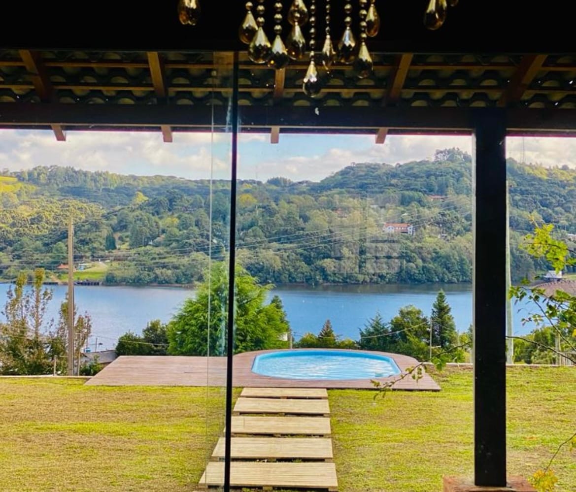 A view of a serene lake is framed by large glass doors, leading to a grassy area. A wooden pathway extends towards a circular pool, surrounded by lush greenery and gentle hills in the background, enhancing the tranquil setting.