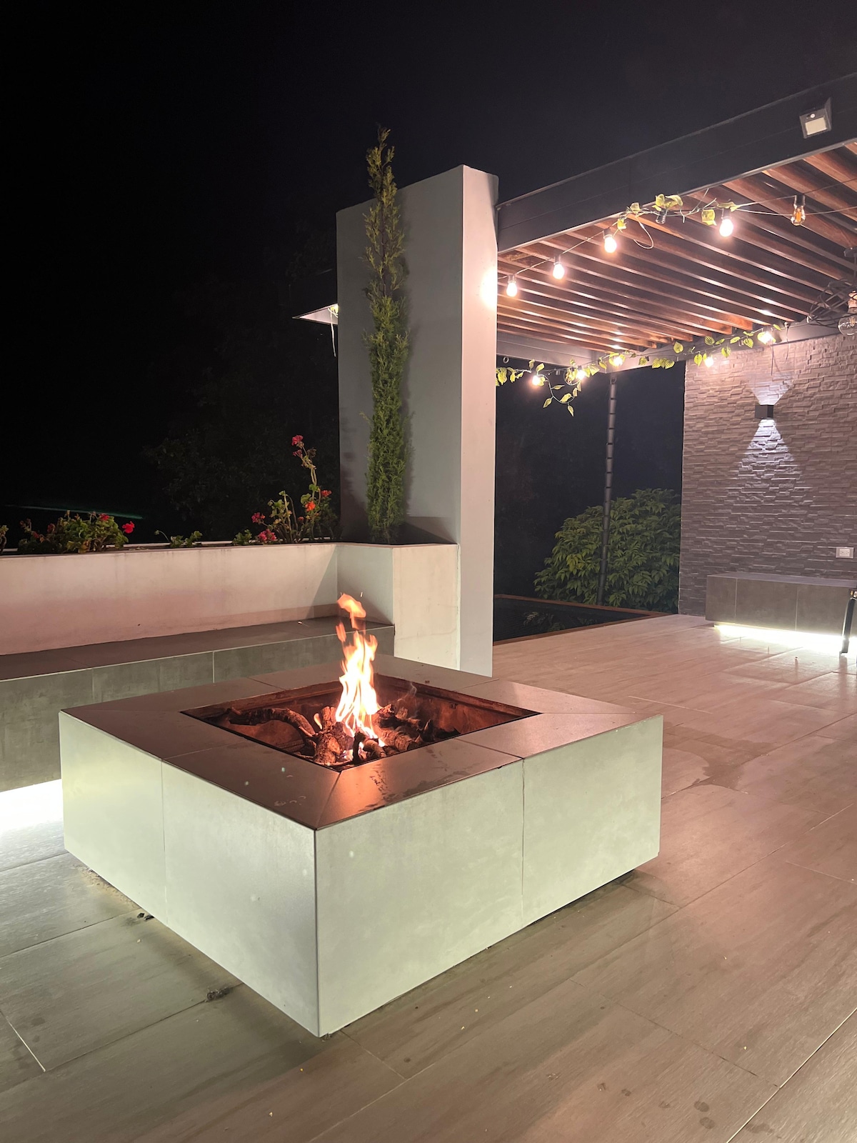 A modern outdoor BBQ and bonfire area features a square fire pit surrounded by sleek, light-colored stone. Soft outdoor lighting creates a warm ambiance, and greenery decorates the space, contributing to a welcoming atmosphere for gathering and relaxation.