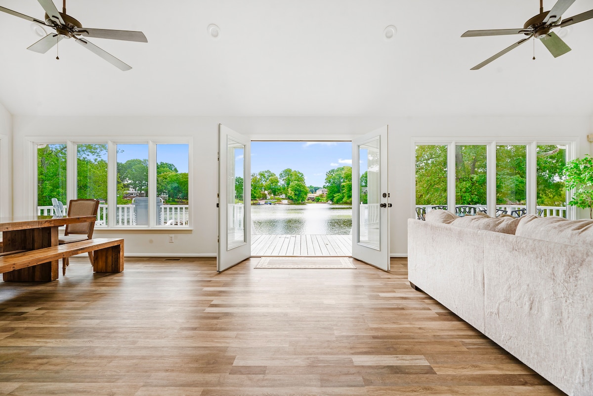 A spacious living area features large glass doors that open to a serene lake view. Natural light fills the room, highlighting the wood flooring. A comfortable sofa and a rustic wooden dining table are positioned to take in the scenic surroundings.