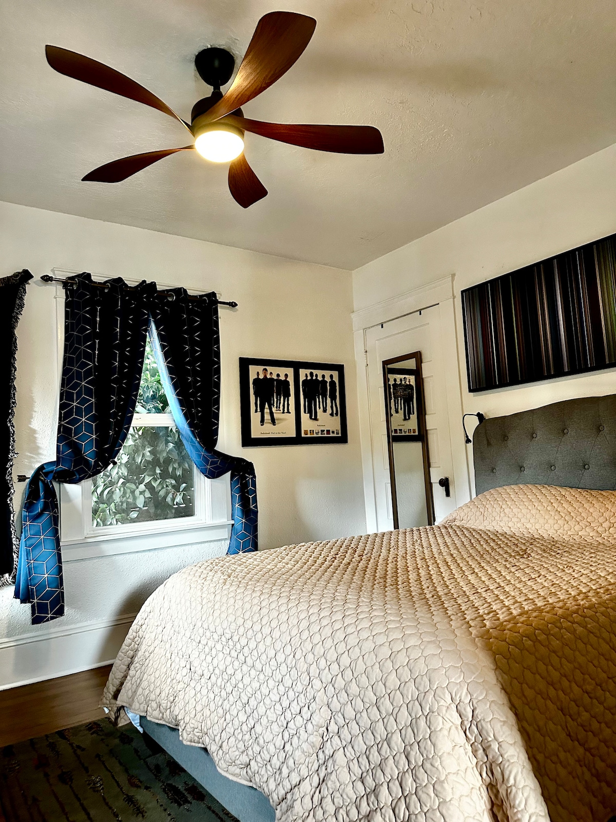 The airy bedroom features a spacious bed covered with a light quilt. A ceiling fan provides a gentle breeze, and framed artwork adorns the walls. Sheer curtains frame a window that invites natural light, creating a comfortable, serene atmosphere.