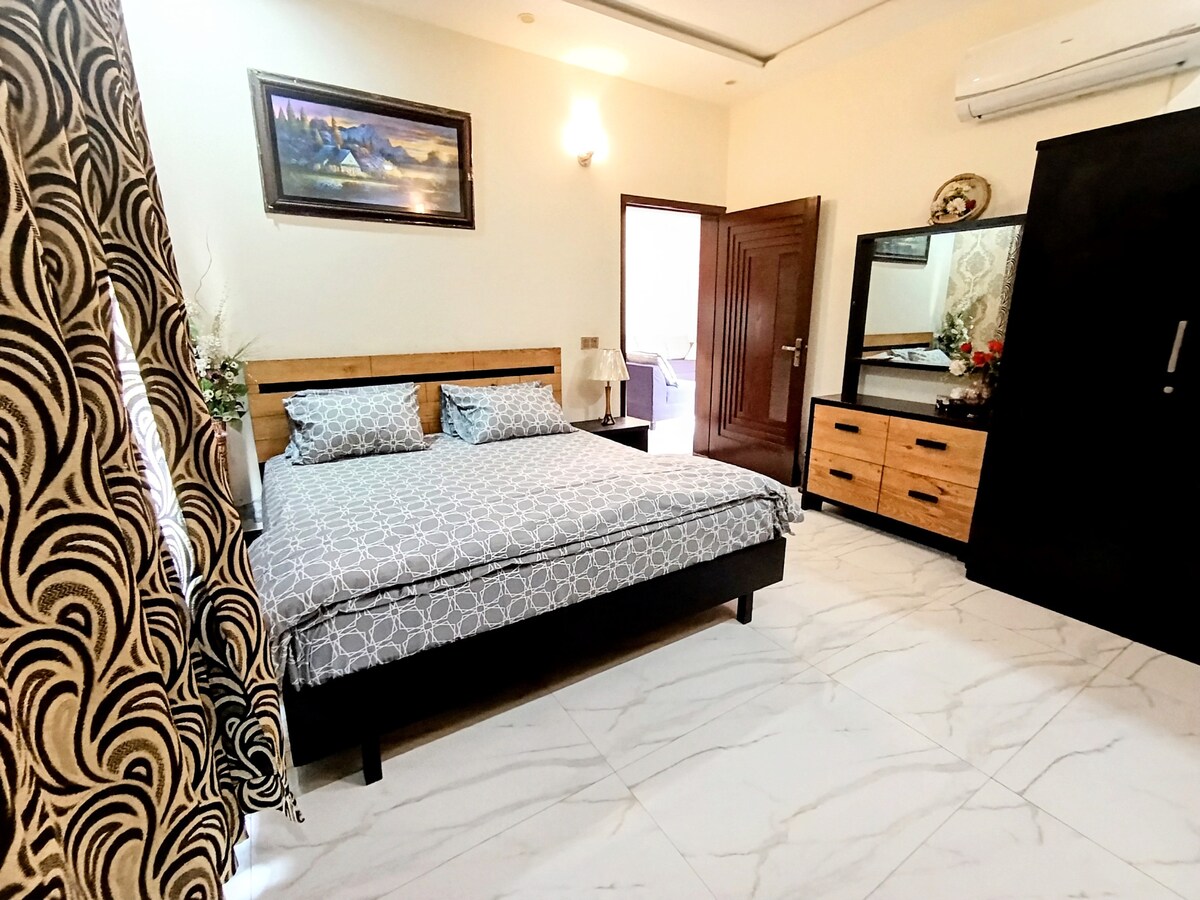 A cozy bedroom features a spacious bed with patterned linens and decorative pillows. Light is provided by a nearby lamp, and a mirror sits atop a wooden dresser, complemented by a stylish wardrobe and artwork on the wall. A window is partially framed by patterned curtains.