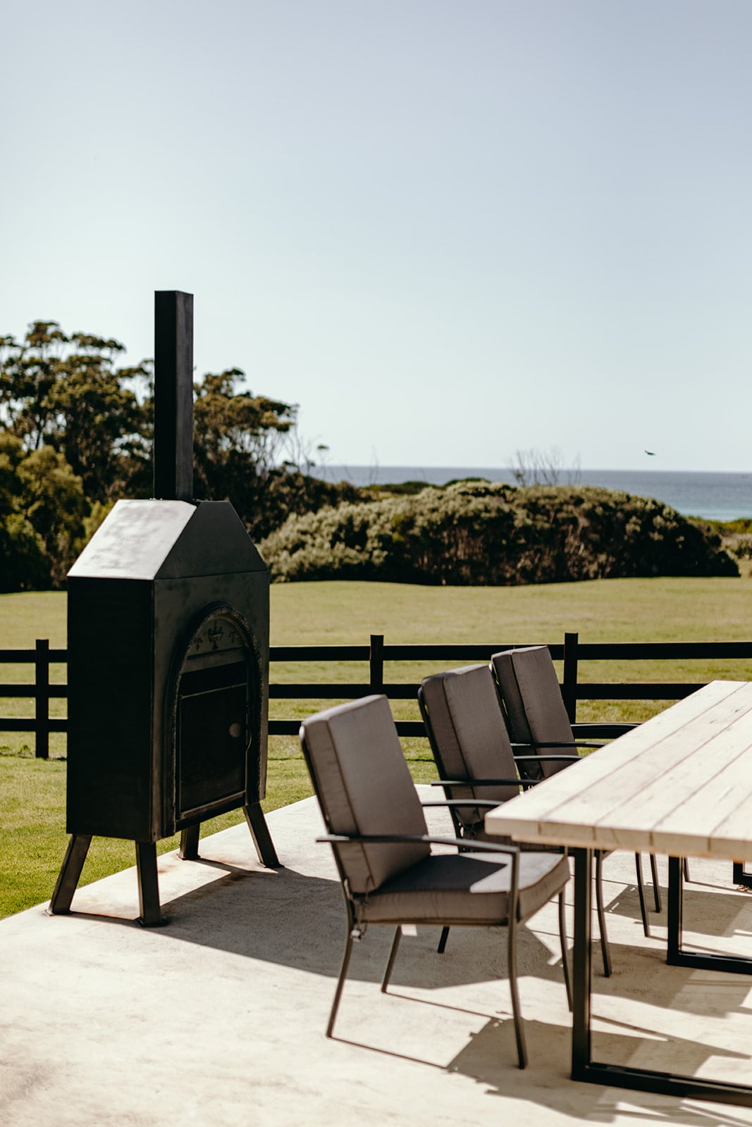 An outdoor dining area features a long table surrounded by several chairs, with a modern barbecue grill positioned nearby. The ocean is visible in the background, beyond a green lawn that gently slopes towards the beach, enhancing the serene setting.