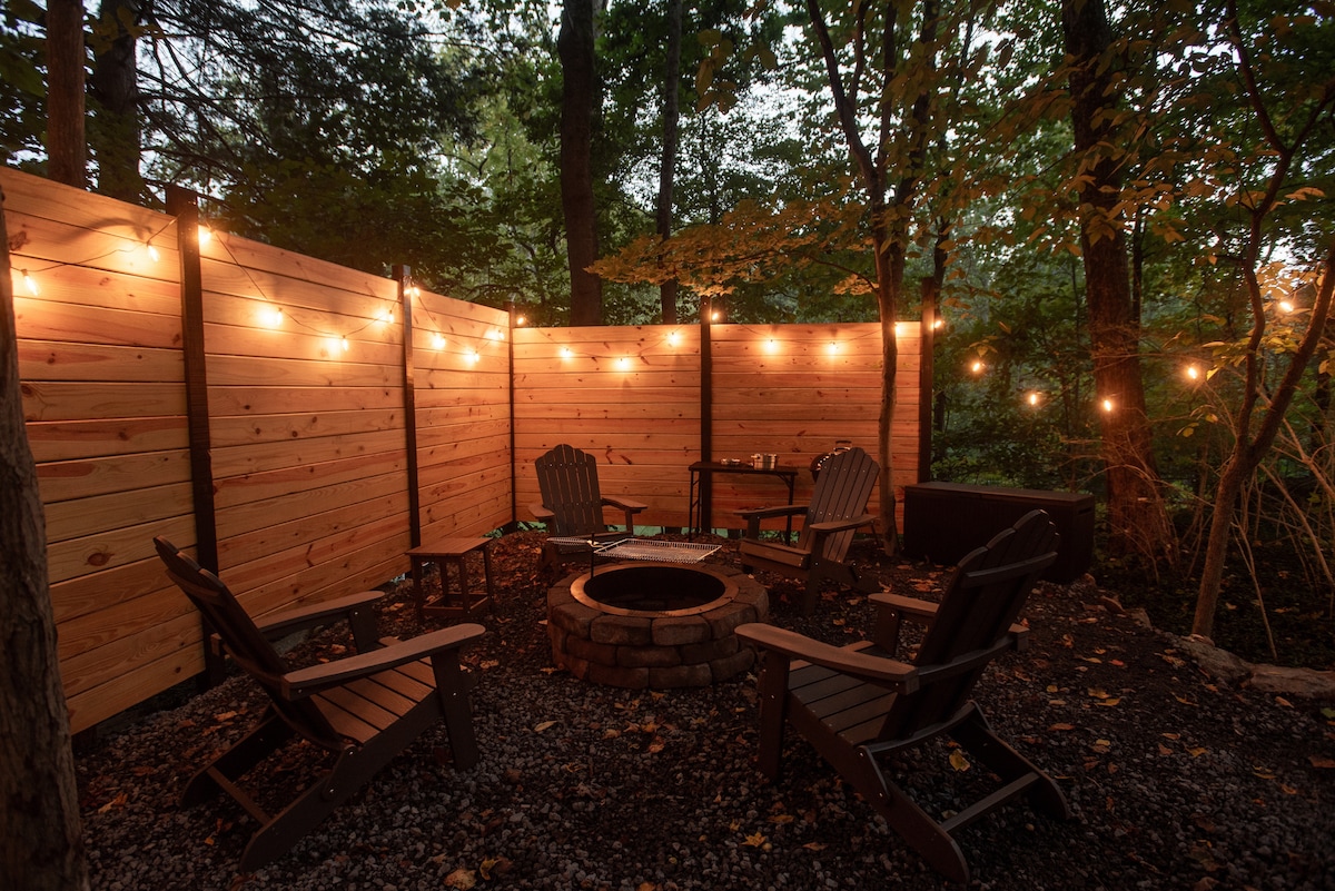 A cozy outdoor seating area is surrounded by illuminated wooden panels, creating a serene atmosphere. Four Adirondack chairs are arranged around a stone fire pit, with string lights hanging above. A small table sits nearby, offering additional space for refreshments or snacks.