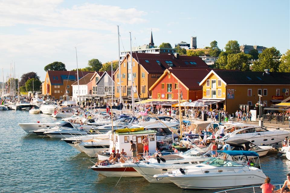 The image displays a vibrant harbor filled with several boats docked close to the shore. Colorful buildings line the waterfront, with people enjoying their time in the area. A clear sky and sunshine create a lively atmosphere around the marina.