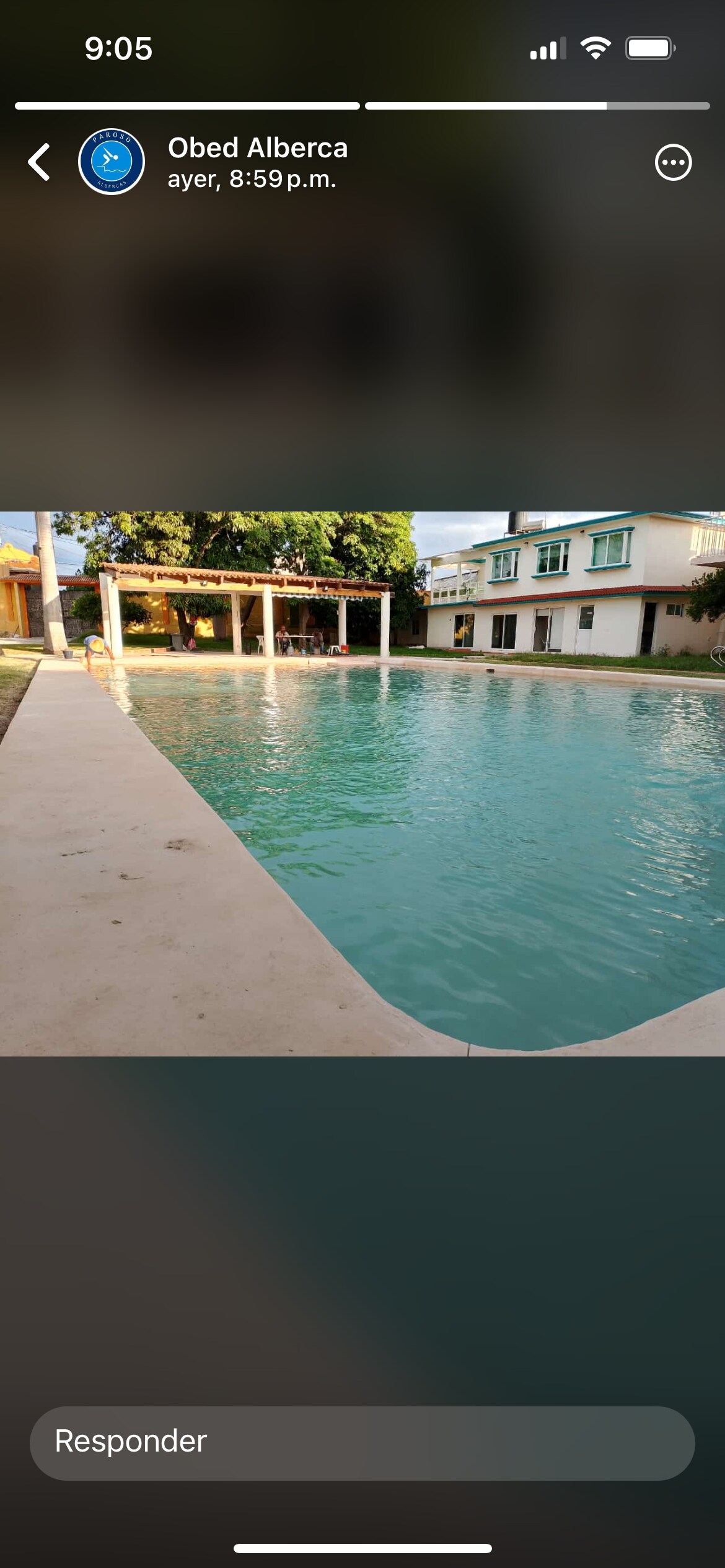 A large swimming pool is framed by smooth concrete edges, reflecting the surrounding greenery. A shaded seating area with a roof is visible next to the pool, providing a space for relaxation. The two-story building can be seen in the background, featuring multiple windows.